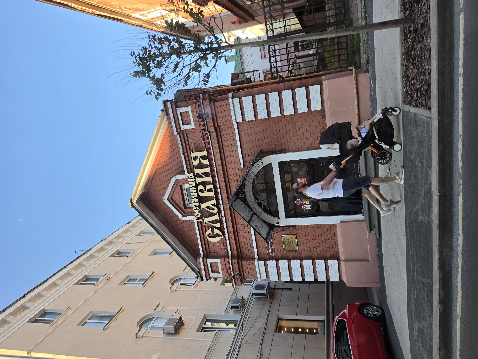 A man, wearing a white t-shirt and dark shorts, pushes a stroller with a young child in it along a sidewalk. The child is wearing a blue shirt and white shoes. The man is walking to the right, and the stroller is facing away from the viewer. Behind them is a red brick building with a sign that says "ГОСТИННИЦА СЛАВИЯ" in gold letters. The building has a green roof and white decorative elements. To the left of the building is a light-colored, multi-story building with windows and air conditioning units. A red car is parked on the street in front of this building. To the right of the brick building is a metal fence and a tree. The sky is clear and blue, with a crescent moon visible. The scene appears to be in an urban setting, likely a city street in Nizhny Novgorod, Russia.
