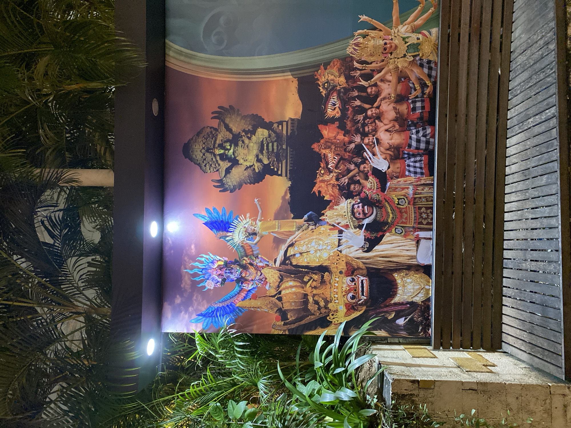The image displays a large mural depicting a Balinese cultural performance, likely a Legong dance or a similar traditional dance, situated in Kuta Selatan, Indonesia. The mural is illuminated by overhead lights, highlighting the vibrant costumes and dramatic masks of the performers. In the foreground, a large, fearsome mask with a red face and white eyes is prominently featured. Behind it, figures in elaborate headdresses and traditional checkered sarongs are visible, some with their hands raised. A central figure, adorned with intricate gold detailing and multiple arms, embodies a divine or mythical being. The backdrop of the mural features a large statue of a winged deity, possibly Garuda, and stylized dragon-like creatures. The mural is mounted on a dark panel above a wooden bench and seating area. Lush green foliage, likely palm trees, surrounds the mural, suggesting an outdoor setting within a tropical environment. The overall scene evokes a sense of cultural richness and artistic expression.