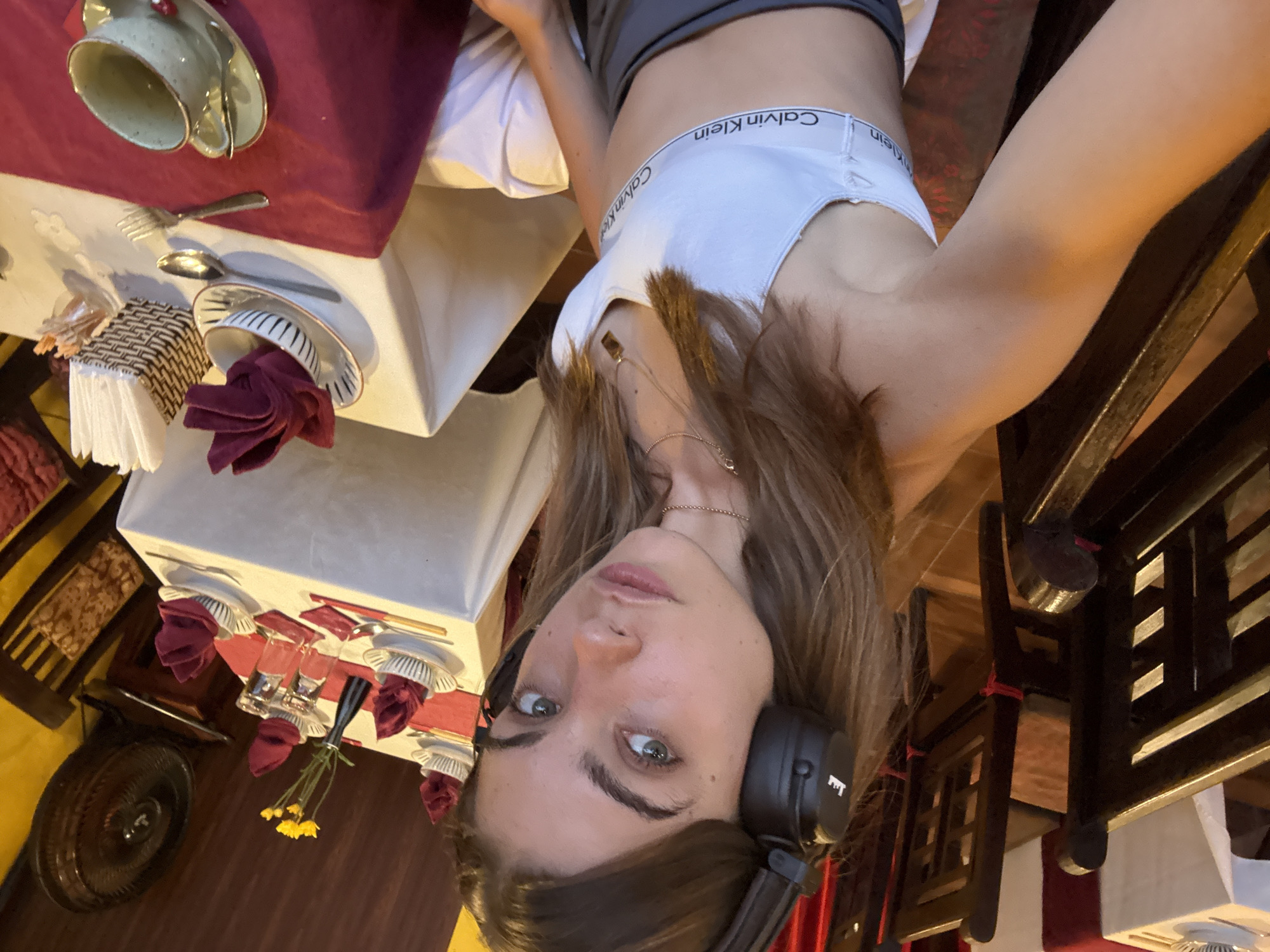 A woman with brown hair, wearing headphones and a Calvin Klein sports bra, is taking a selfie in a dining area. She is positioned at an angle relative to the camera. The background shows several tables set for dining, with plates, glasses, napkins, and cutlery arranged on white tablecloths with maroon accents. A fan is visible in the lower left corner, and chairs with a dark wood finish are around the tables. The scene suggests a restaurant setting in Hội An Ward, Vietnam.