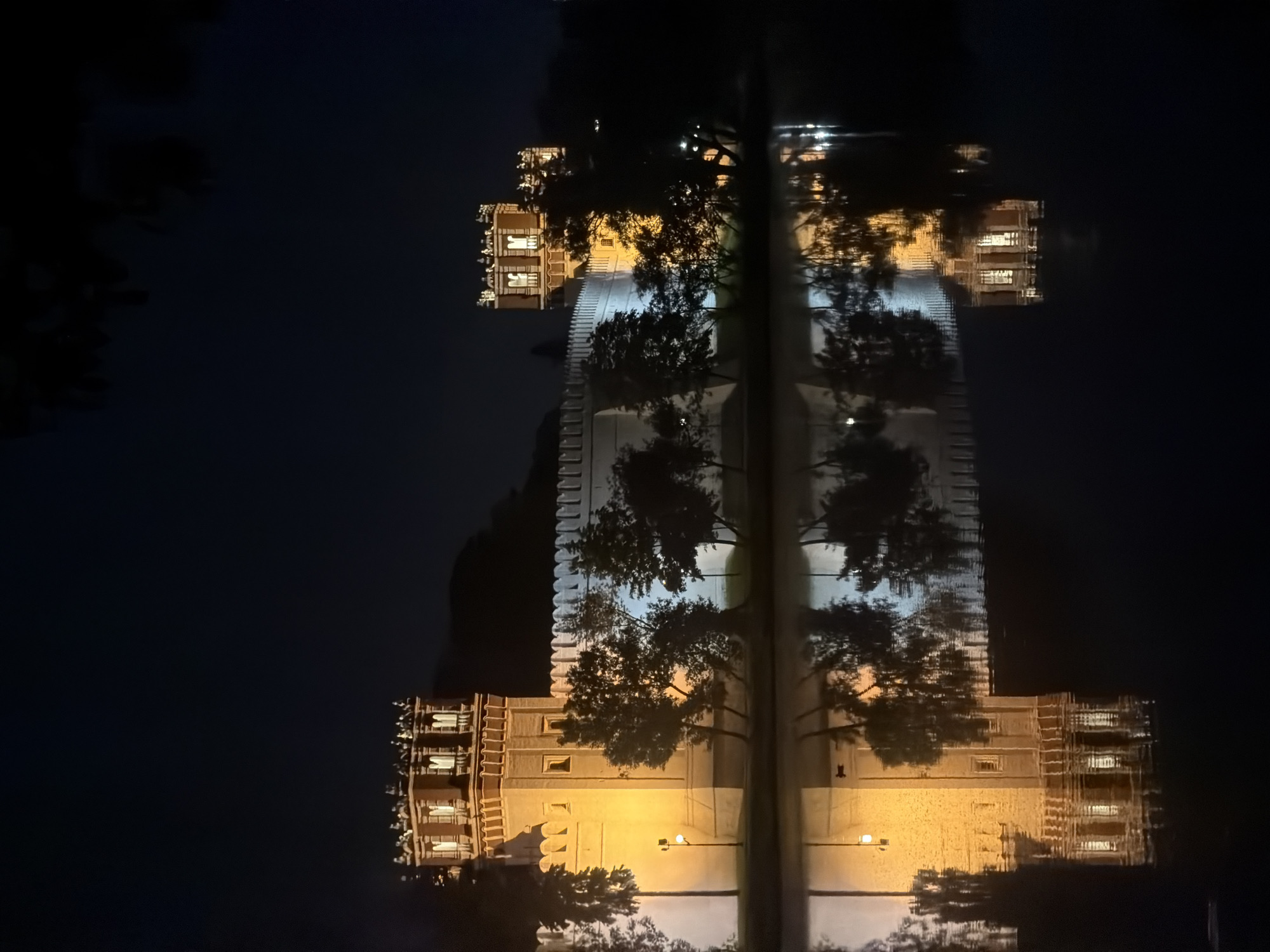 The image displays a majestic architectural structure at night, dramatically illuminated and perfectly reflected in calm water. The main subject is a large, grand building, likely a fortress, palace, or a historical complex, characterized by its imposing size and distinctive features. Its light-colored facade, possibly stone or stucco, is bathed in warm, yellow-orange artificial light, which accentuates its architectural details, including crenellated battlements along its upper edges and multiple windows, some of which glow with interior light.
Dark silhouettes of trees are visible in the foreground and partially obscure parts of the building, both the actual structure and its reflection. The still surface of what appears to be a river or canal acts as a pristine mirror, creating an almost perfect, inverted reflection of the illuminated building and the trees below. The sky is dark, indicating that the photograph was taken at night. No human figures or activities are visible, emphasizing the tranquil and serene atmosphere of the scene. The clear reflection suggests calm weather conditions. Based on the provided context, the building is located in Moscow, Russia, and its style is reminiscent of Russian historical architecture. There is no visible text in the image.