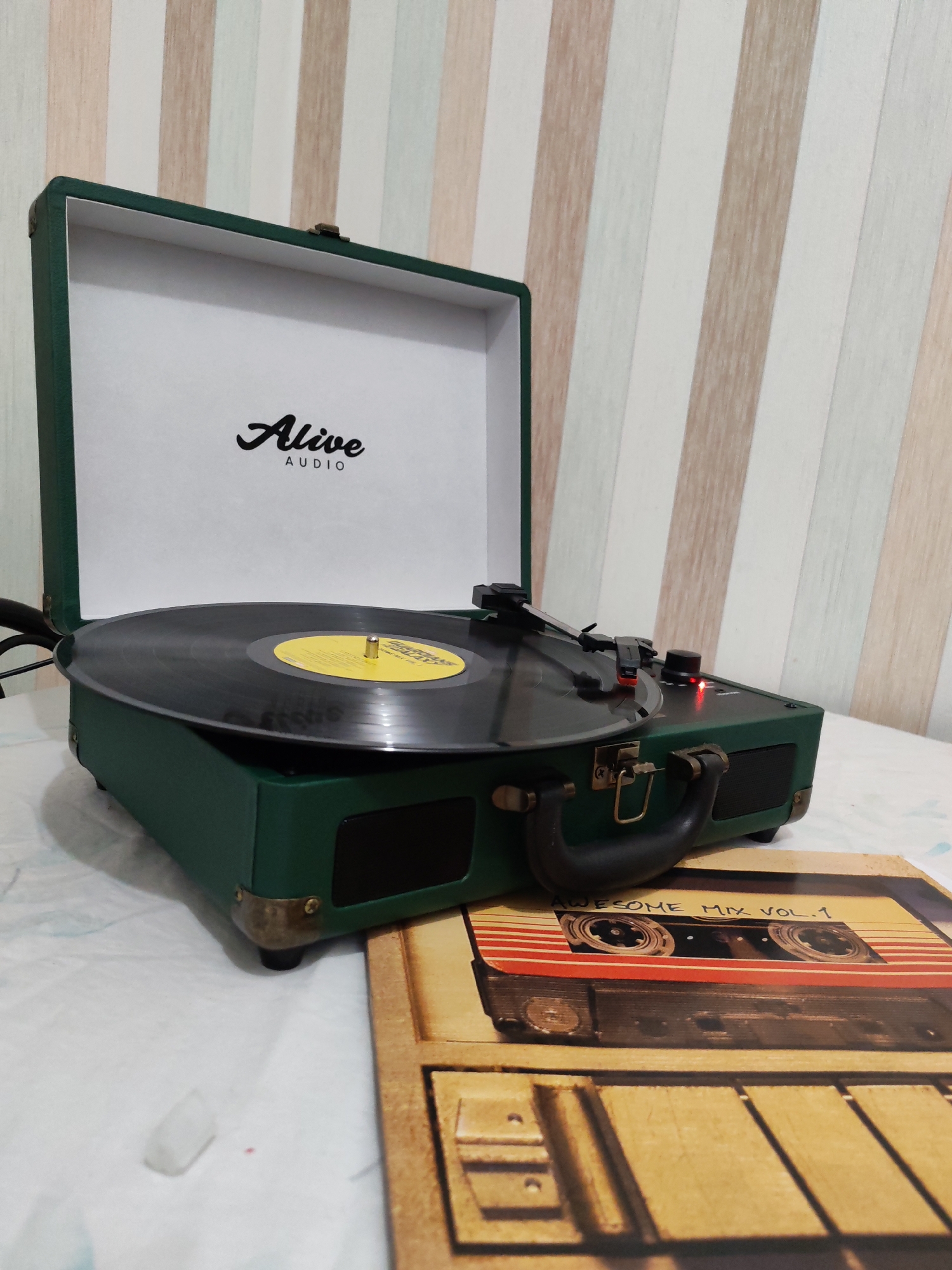 A dark green, portable suitcase-style record player is prominently displayed in an indoor setting in Izhevsk, Russia. The record player is open, revealing its white interior lid with the black "Alive AUDIO" logo printed centrally. A black vinyl record with a yellow center label rests on the turntable, and its tonearm is positioned over the record, suggesting playback or readiness to play. A small red LED indicator light is visible on the right side of the player's control panel. The record player features brass-colored latches and a black carrying handle on its front, along with speaker grilles.
It is placed on a light-colored surface, possibly a tablecloth or bedspread. To the right and slightly in front of the record player lies an album cover or sleeve. This cover features a graphic design of an old-fashioned audio cassette tape, with the text "AWESOME MIX VOL. 1" clearly visible on its label. A small, irregularly shaped white object is visible on the light surface in the bottom left corner. The background consists of a wall covered in vertically striped wallpaper, featuring alternating bands of light brown, white, and a pale green or blue color. The scene is brightly lit, indicating daylight or good indoor lighting.