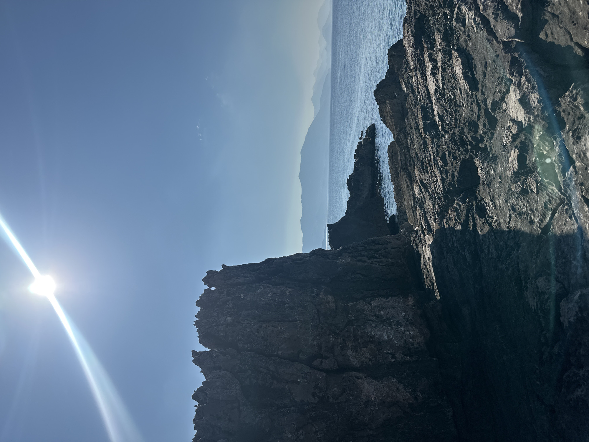 A rugged, dark rock formation dominates the left foreground of this coastal scene in Seoca, Montenegro. The rock face is textured and uneven, with sharp, jagged peaks along its top edge. To the right of the formation, the calm, blue sea stretches towards a distant horizon. The water is clear and reflects the bright sunlight. Faint outlines of layered, hazy mountains are visible in the far distance, appearing as soft silhouettes against the pale blue sky. The sky itself is mostly clear, with the sun positioned in the upper left quadrant, casting a bright glare and lens flare across the image. The rocky ground in the immediate foreground is also dark and uneven, with visible cracks and crevices. Shadows are cast across the rocks, indicating a strong light source. The overall impression is one of a wild and natural coastal landscape.