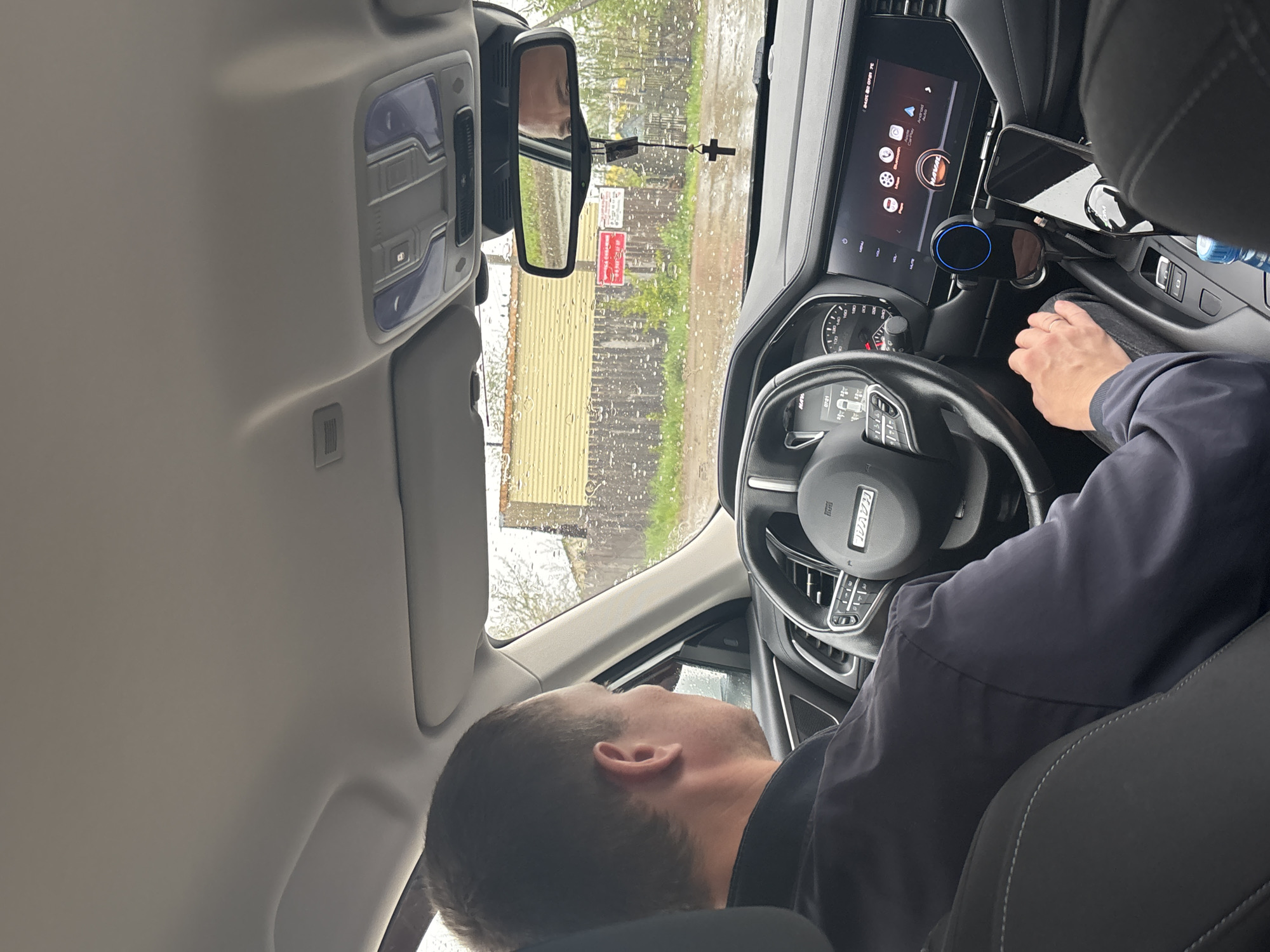 The image captures an interior view from the passenger seat of a vehicle, likely a car, in what appears to be a rainy or wet environment. The driver is visible, although their face is mostly obscured from this angle. Their hand is on the steering wheel, which features the Haval logo, indicating the vehicle's brand. The interior is modern, featuring a digital display screen on the dashboard. A phone is mounted on a holder. The rearview mirror shows an external view with what appears to be a building with yellow corrugated siding and road signs. Considering the specification of the location, it is probable that the scene is in Chaikovsky, Russia. The presence of rain streaks on the windshield suggests ongoing or recent precipitation.