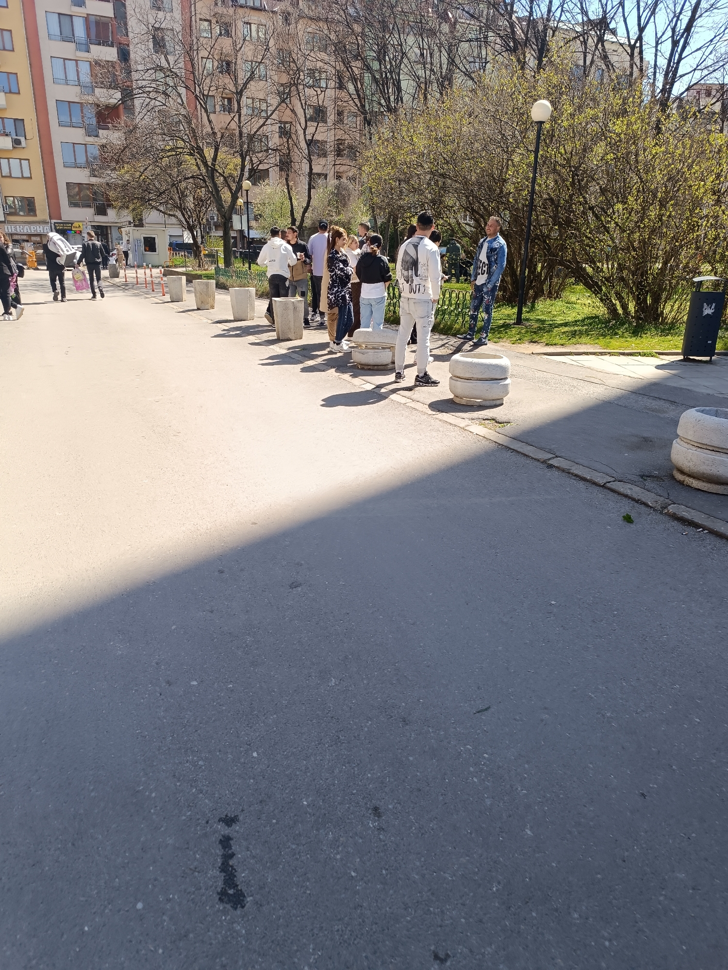 The image shows a group of people standing in a line on a street in Sofia, Bulgaria. The queue is lined with concrete barriers. There is a trash can and some green bushes. In the background are several buildings. The scene is bathed in sunlight. The image was taken during the day.