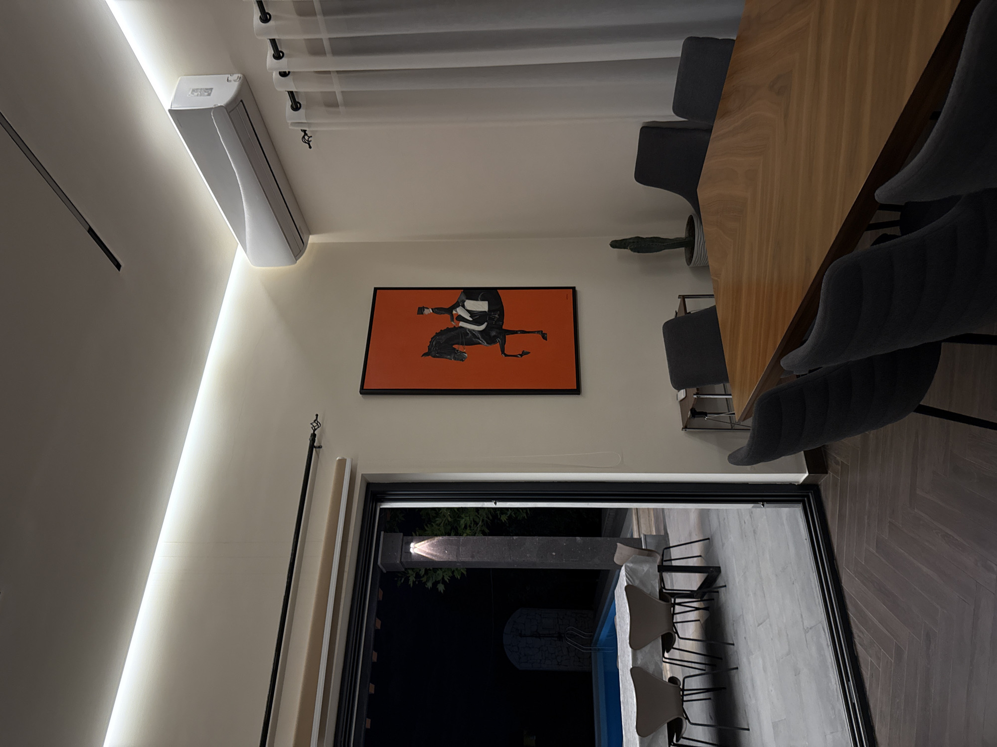 The image shows the interior of a modern space, likely a dining room or living area. The walls are painted in a neutral tone, and an LED light strip illuminates the left side of the frame. A rectangular piece of art hangs on the wall, featuring a stylized depiction of a horse and rider in black against an orange background, framed in black. To the right of the artwork is a dining table, made of light-colored wood, surrounded by chairs upholstered in dark gray fabric. The backs of the chairs are curved for comfort.
A large, sliding glass door provides a view to an outdoor area with a swimming pool. Several chairs can be seen through the open door, and they appear to be the same style as the ones around the dining table. The outdoor area is dimly lit, suggesting it's either nighttime or a covered patio.
Mounted high on the wall near the ceiling is an air conditioning unit. White sheer curtains hang from a rod above the unit. A few green plants are placed near the chairs, adding a touch of nature to the interior. The flooring inside is a dark herringbone pattern. The scene could be located in Yerevan, Armenia, though there are no explicit visual indicators in the image.