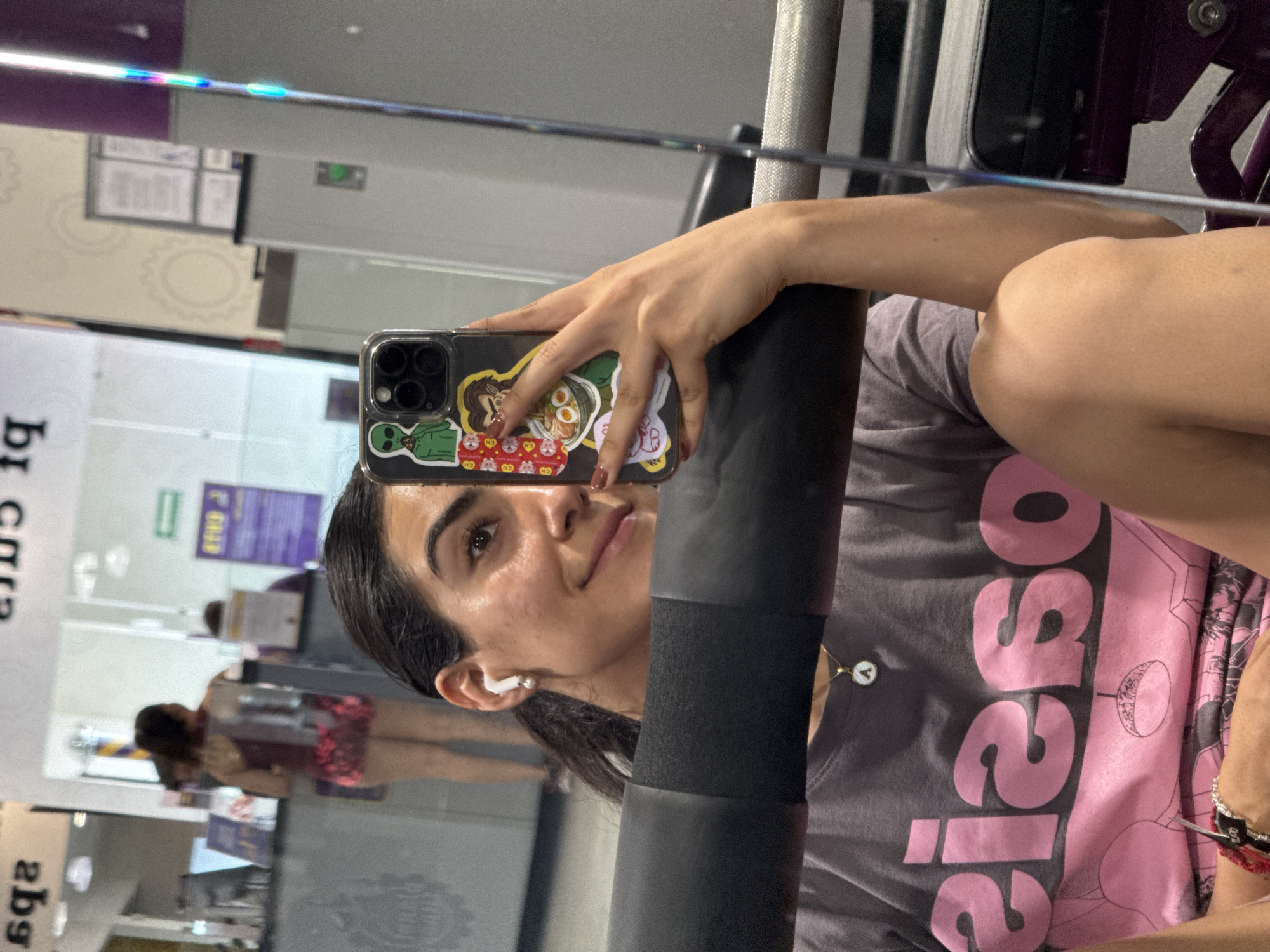 A woman takes a selfie in a mirror at a Planet Fitness gym. She is wearing a grey t-shirt with pink writing and has a phone case decorated with stickers. The gym equipment is visible in the mirror, including a weight machine. Other people are also visible in the mirror, including one individual near a service desk. The location appears to be a Planet Fitness in Puebla, Mexico, based on visual clues such as signage and interior design consistent with the brand's aesthetic.