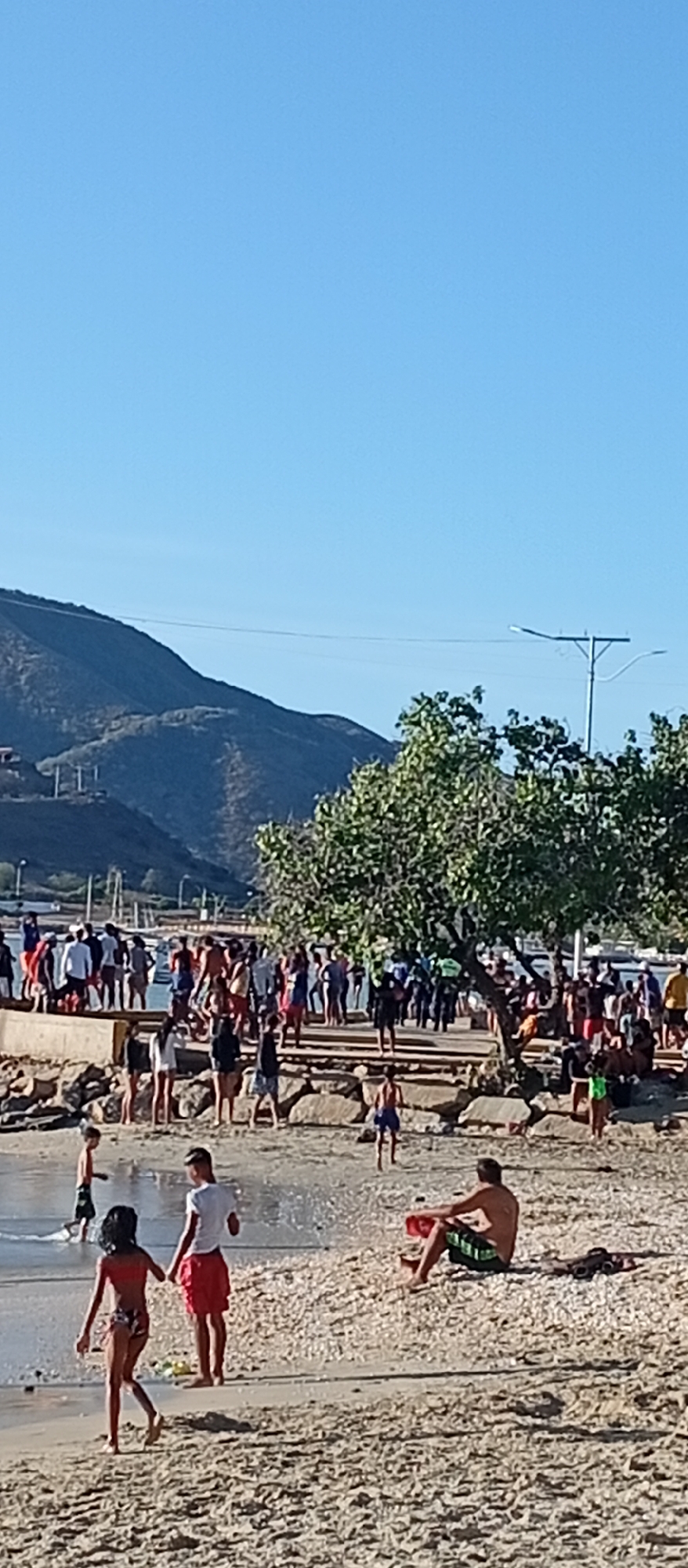The image depicts a crowded beach scene in Juan Griego, Venezuela. A group of people are congregated on a concrete pier or walkway extending into the water. Individuals are also standing and walking on the beach, some in the water's edge. A few are sitting on the sand. In the background, there are trees and mountains under a clear blue sky. There is also a lamppost in the background. The beach surface transitions from sand in the foreground to pebbles and rocks closer to the water. The overall impression is a busy beach day.