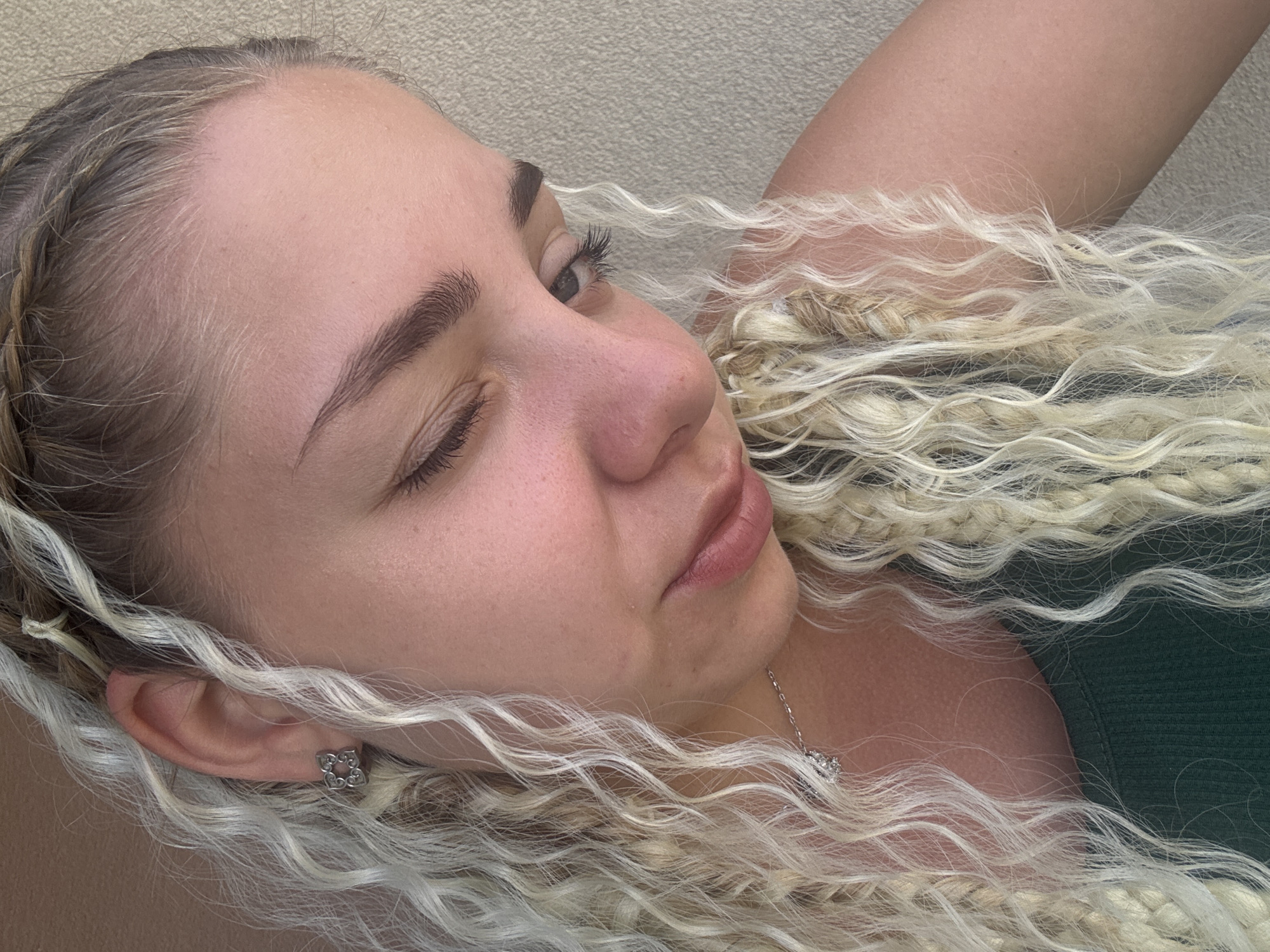 The image shows a close-up of a young woman with blonde, curly hair, some of which is braided. She is wearing a green top and a silver necklace with a small pendant. She also has a silver earring shaped like a flower in her left ear. The woman is looking down with her eyes partially closed. The background is a plain, textured wall, possibly an exterior wall of a building. The lighting suggests it is daytime. The image does not contain any specific information or landmarks that would allow for its location to be identified in Nesebar, Bulgaria.