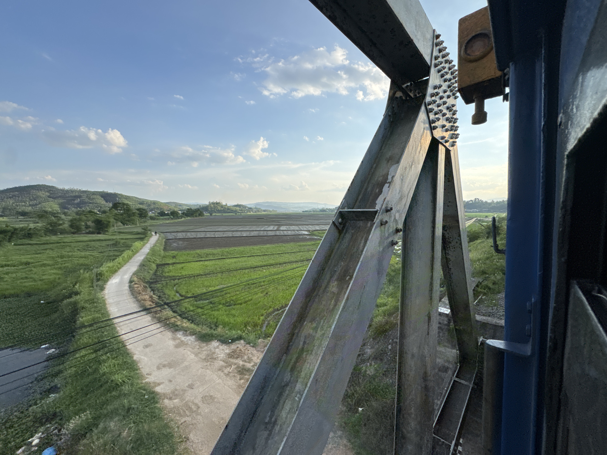 An exterior, low-angle, wide shot captures a view from a train as it crosses a bridge, looking out over a rural landscape in Chí Thạnh, Vietnam. The foreground is dominated by the imposing steel structure of the bridge, with its beams and rivets clearly visible. To the right, a section of the train's blue exterior is also in frame. Beyond the bridge, a winding dirt road curves through vibrant green rice paddies and fields. In the distance, rolling hills under a bright blue sky dotted with fluffy white clouds complete the serene, pastoral scene. There are no people visible in the image.