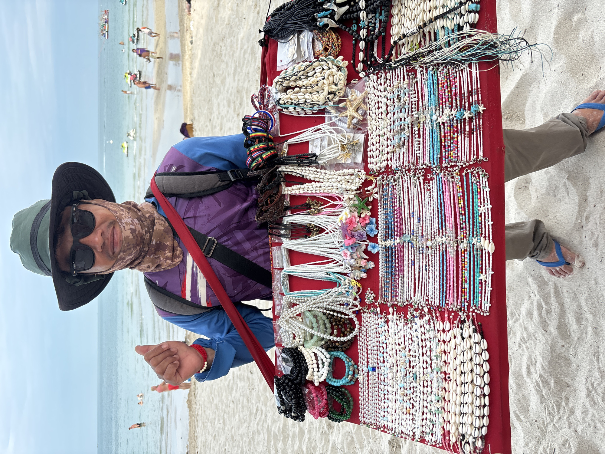 The image shows a man with a portable jewelry stall on a beach. He wears a hat, sunglasses, and a patterned face covering, a blue and purple shirt, and khaki pants. He stands behind a red table laden with necklaces and bracelets made of beads, shells, and other materials. The stall includes necklaces featuring starfish and other sea-themed designs. A red strap crosses his body, supporting the table. The beach stretches behind him, with people visible in the distance along the shoreline and in the water. The sand is light-colored, and the sea and sky are a light blue. The location is Baan Chaweng, Thailand.