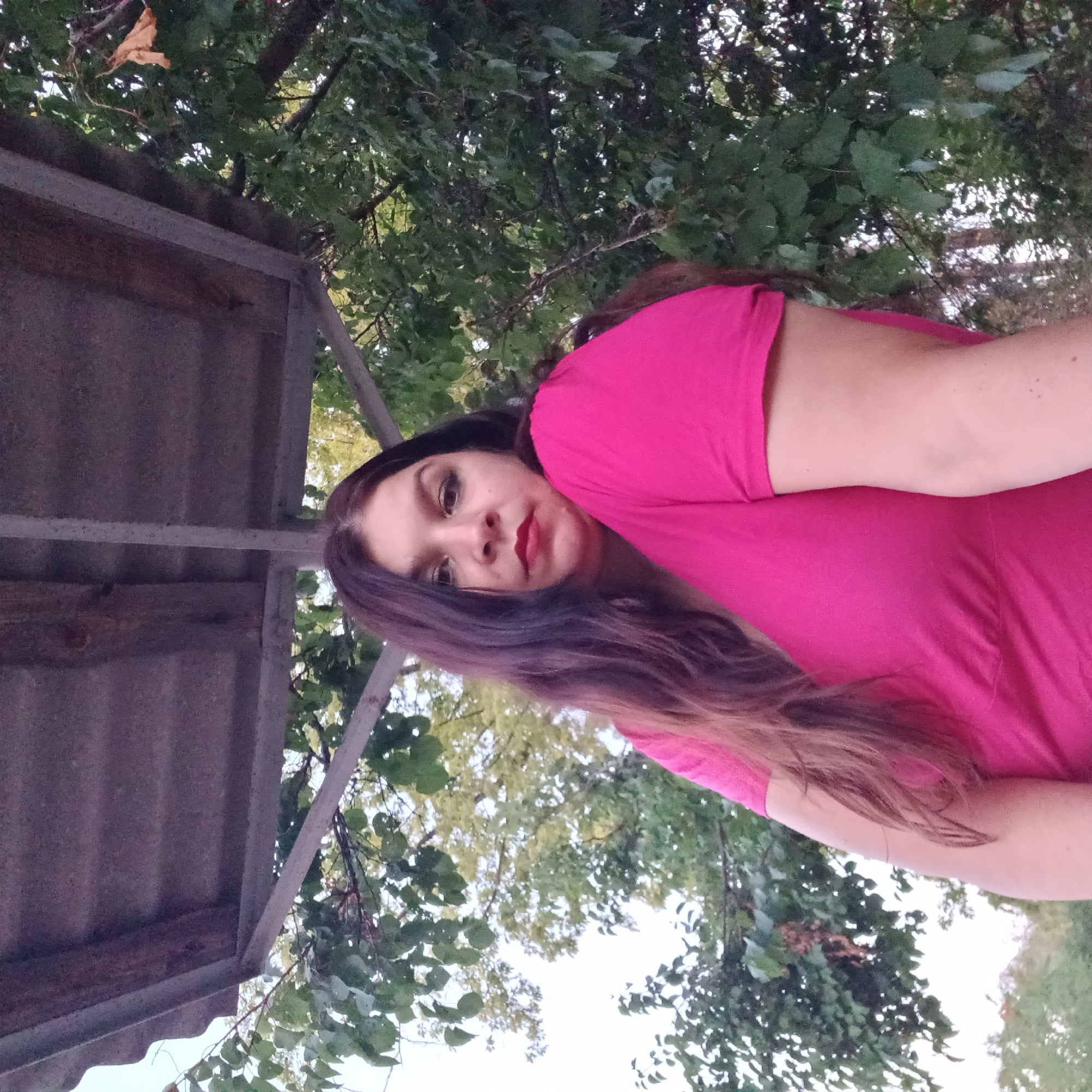 A woman with long, dark hair and red lipstick is looking at the camera in an outdoor setting. She is wearing a bright pink t-shirt. Behind her, there are green trees and a wooden structure overhead. The lighting suggests it is daytime. The image does not provide specific details to determine a location within Berezhynka, Ukraine.