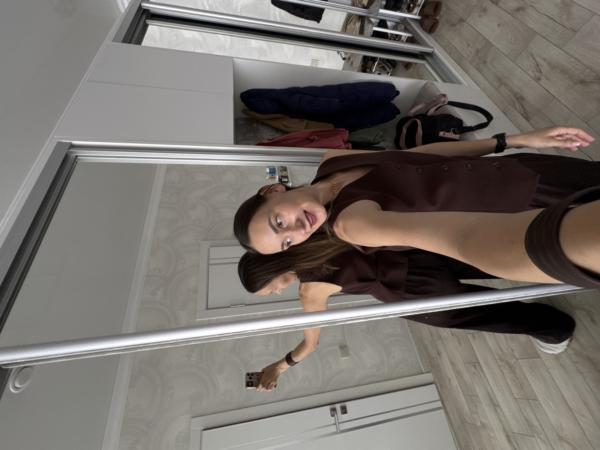 A woman is taking a mirror selfie in what appears to be a bedroom or dressing area, likely in Sochi, Russia. She is shown from the chest up, looking directly at the camera with a slight smile and an engaging expression, her mouth slightly open.
She wears a dark brown sleeveless top or vest with buttons, paired with matching dark brown pants. A black watch is visible on her left wrist, and a gold choker necklace with the word "DIOR" spelled out in letters is around her neck. Her reflection in the lower part of the mirror shows her holding a gold-colored smartphone with her right hand to capture the image. White sneakers are visible on her feet in the reflection.
The setting is an indoor space dominated by a large white sliding wardrobe with mirrored doors. Inside the wardrobe, shelves hold folded clothes, including a dark blue puffer jacket and other garments. A black shoulder bag with pink accents is also visible on a shelf. The room features light-colored walls with a subtle, repeating circular or scale-like pattern wallpaper and a light-toned wooden floor. White door frames, trim, and what appears to be a light switch are also visible on the walls.