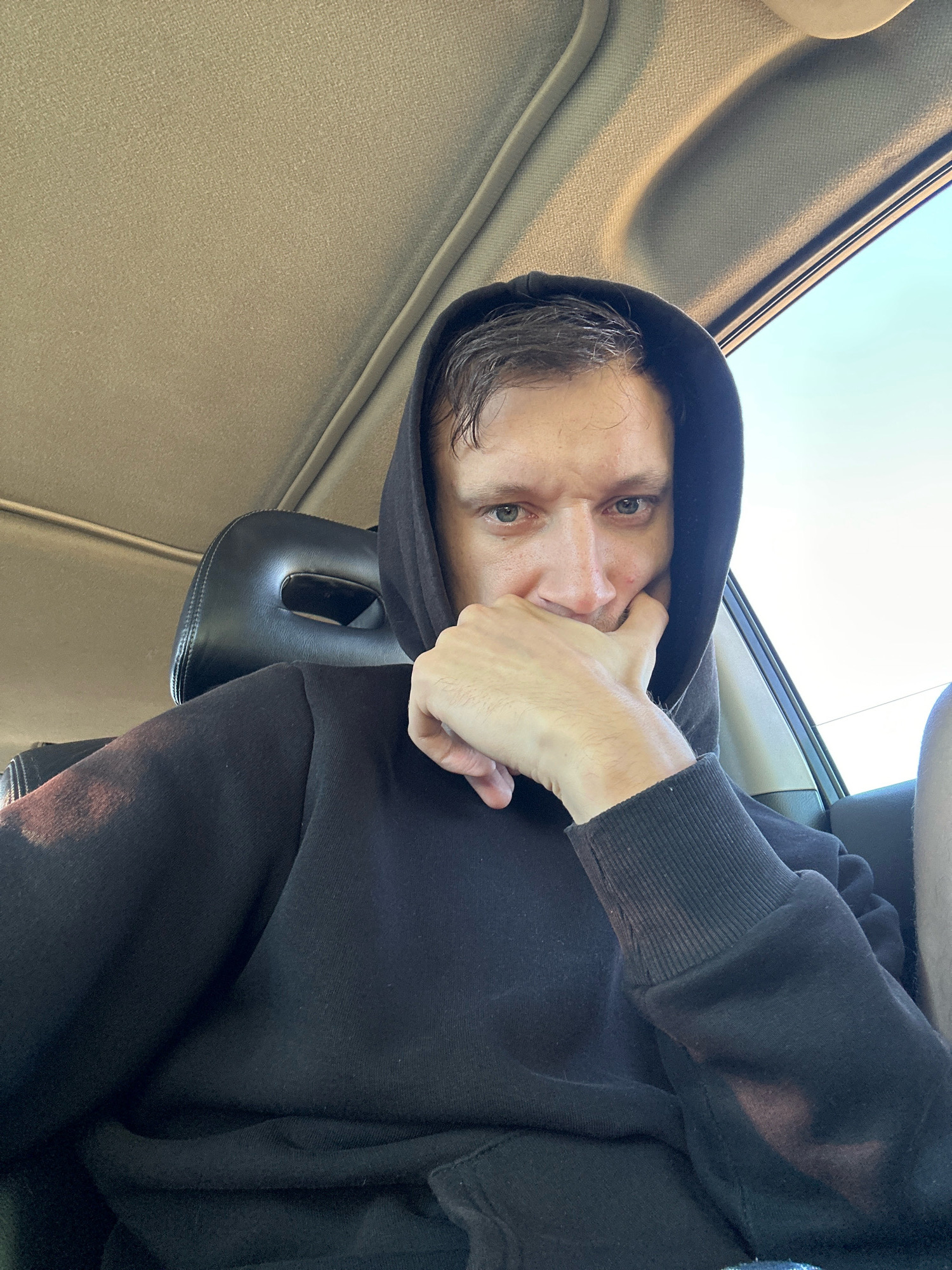 The image shows a man sitting in a car, wearing a black hoodie and resting his chin on his hand. The interior of the car is visible, including the headrest and the roof lining. The exterior of the car is also visible through the window, showing a bright sky. The scene is set in the city of Makiv, Ukraine.