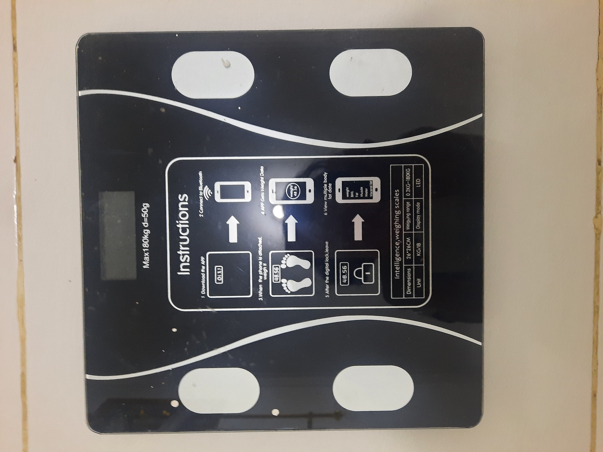 An overhead, eye-level, slightly tilted, medium shot captures a black digital scale placed on a tiled wall. The scale, featuring a white curved line design, has a digital display at the top showing "Max180kg d=50g". Below, a section labeled "Instructions" provides a numbered, step-by-step guide with icons and text. The instructions detail downloading an app, connecting to Bluetooth, weighing oneself, and viewing body fat data on a smartphone. The scale itself has two white oval-shaped footpads and a prominent white circular button. At the bottom of the scale, a specification table lists "Dimensions 26*26CM", "Weighting range 0.2KG-180KG", "Unit KG/IB", and "Display mode LED". The background consists of a beige tiled wall with some visible dirt and debris, suggesting a domestic or commercial environment in Lhokseumawe, Indonesia.