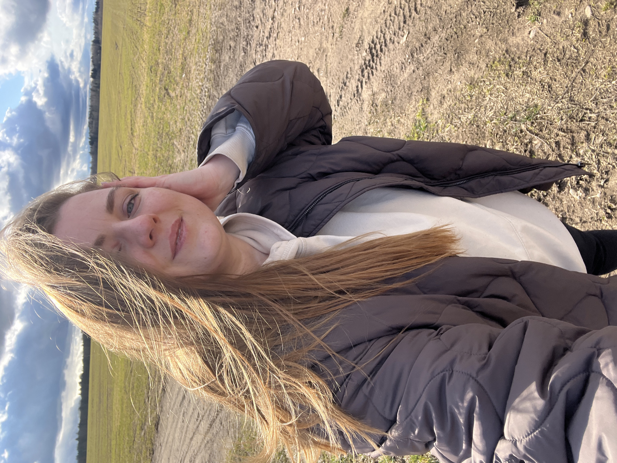 A woman poses for a selfie in an outdoor setting that appears to be a field, possibly in Ямуга, Russia. The woman is wearing a brown quilted jacket over a light-colored hoodie. She has long blonde hair and is touching her face with her hand. The background consists of a field with some vegetation and a cloudy sky. The ground appears to be dirt or gravel.
