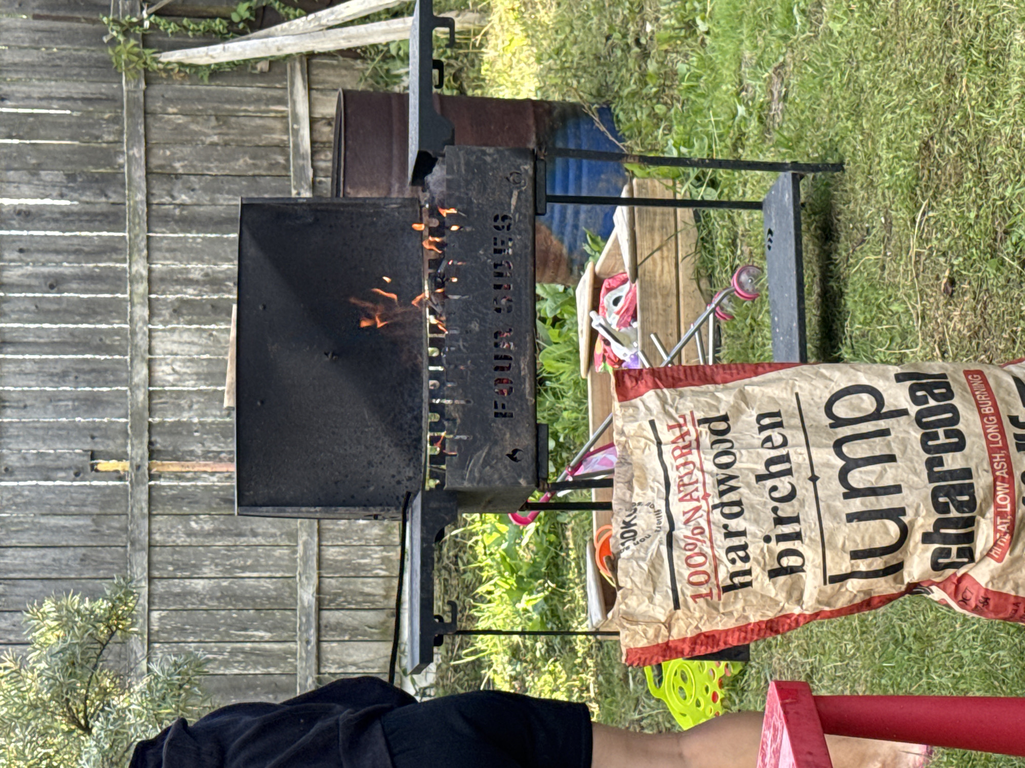 The image depicts an outdoor scene, likely a backyard, with a portable charcoal grill in the foreground. Flames are visible inside the grill, indicating it is in use. To the left of the grill, a large bag of "100% NATURAL hardwood birchen lump charcoal" is prominently displayed, with text indicating "HIGH HEAT, LOW ASH, LONG BURNING". Behind the grill, a weathered wooden fence forms the backdrop. To the right of the grill, a dark-colored metal barrel, possibly a repurposed oil drum, stands partially visible. In the immediate foreground, a person's legs and lower torso are visible, wearing dark shorts and a dark hooded jacket, suggesting they are standing near the grill. Various gardening tools and children's toys, including a bright green plastic item and parts of a stroller, are scattered on the grass around the grill and the charcoal bag. The ground is covered in green grass, with some patches of less maintained vegetation. The setting appears to be a residential area, possibly in the town of Подолино Ст, Russia, given the context of the request.