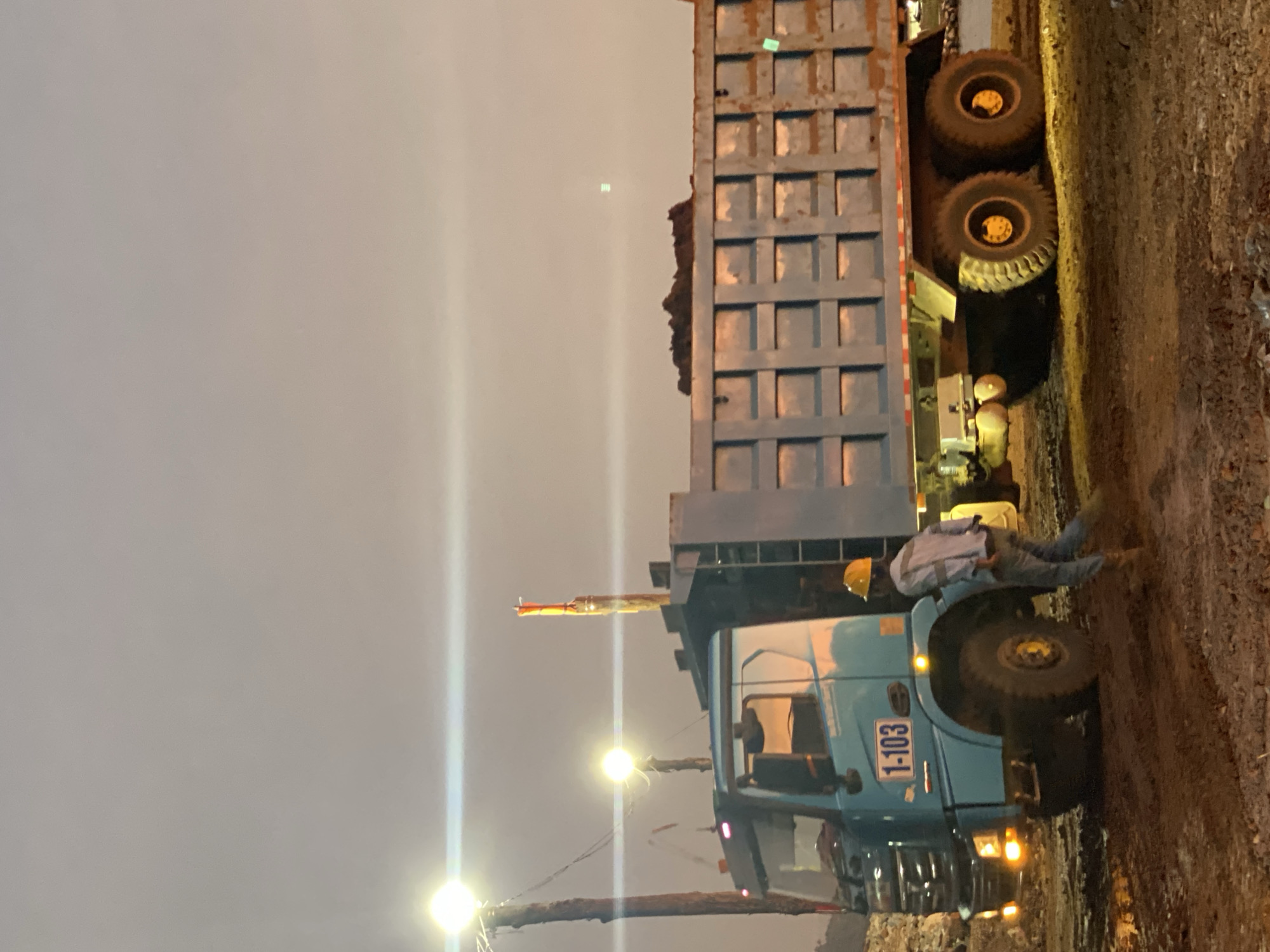 In the city of Sambalagi, Indonesia, at dusk, a large blue dump truck is parked on a muddy construction site. A worker wearing a yellow hard hat and a light-colored long-sleeved shirt and blue pants is walking away from the truck. The truck bed is filled with dark material, possibly soil or rubble. Two large lights on poles are illuminating the area, casting a glow on the truck and the surrounding ground. The sky is overcast and the overall atmosphere is dim, suggesting that work is continuing into the evening. The truck has the number "1-103" visible on its side. The ground is uneven and wet, indicating recent activity or rainfall.