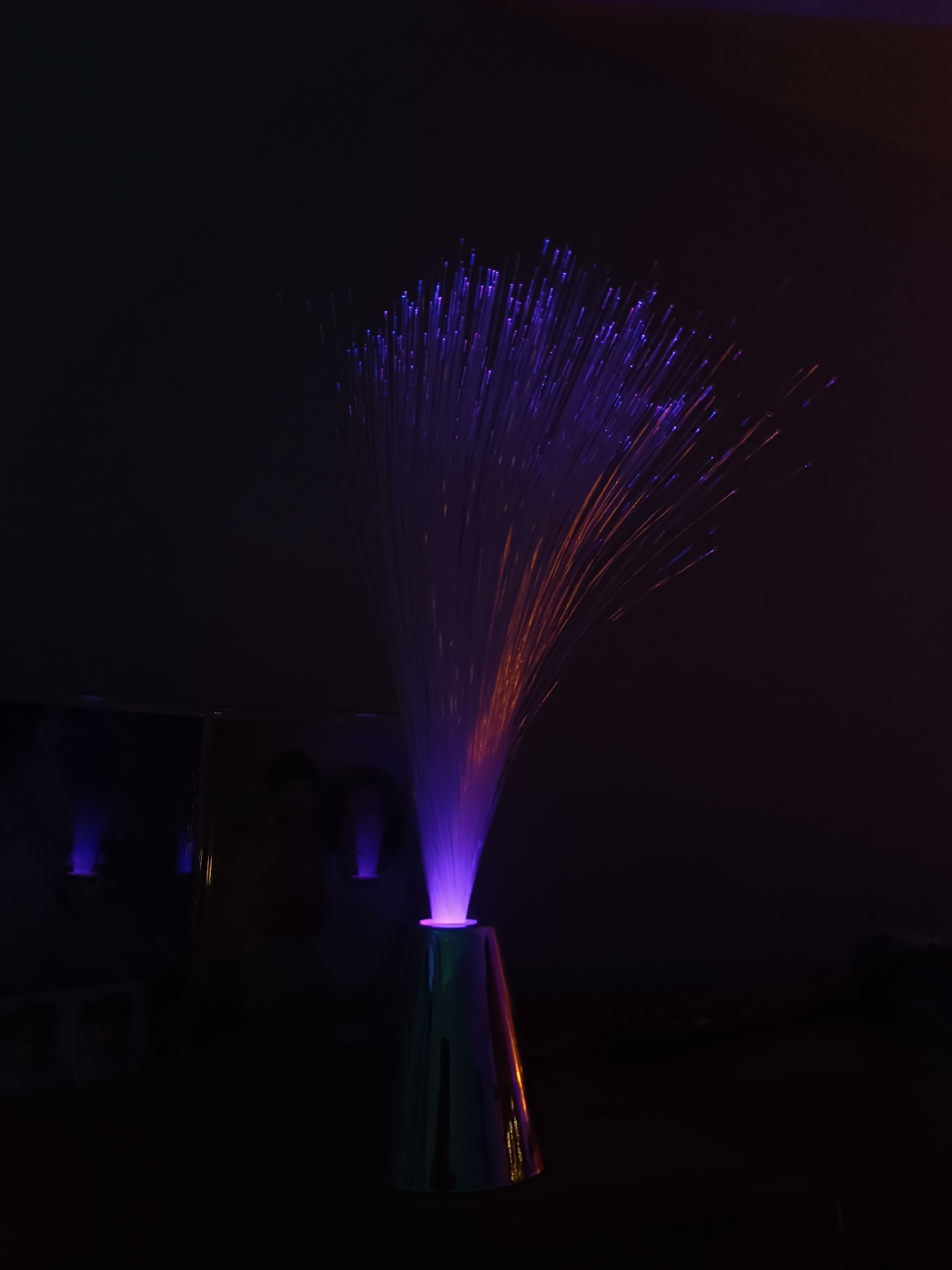 The image shows a fiber optic lamp in a dark environment. The lamp is cone-shaped and the base appears to be silver or reflective. The fibers emerging from the top are illuminated with purple and orange light. In the background, there is a blurry shape that might be another object or a reflection. The lighting is dim, suggesting the lamp is the primary light source. The image does not contain any people, actions, or interactions, and does not provide information regarding the city of Cherkovitsa, Bulgaria.