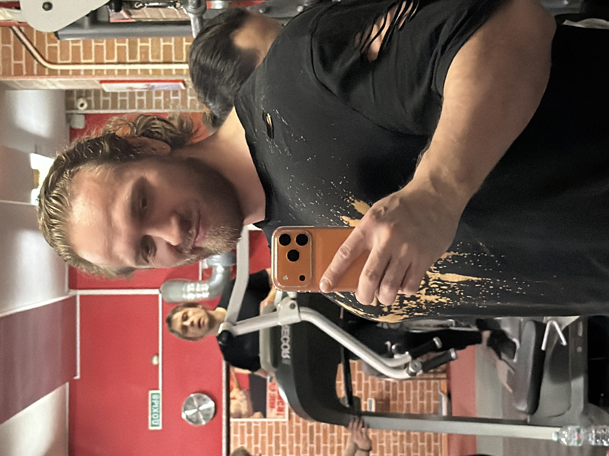 The image displays an interior view of a gym or fitness center located in Saint Petersburg, Russia.
In the foreground, a Caucasian male, estimated to be between 30 and 45 years old, with light brown, wavy, shoulder-length hair pulled back and a light beard, is taking a selfie. He is wearing a black t-shirt adorned with gold paint splatters on the front and distinct vertical rips on the left sleeve and a horizontal rip on the left shoulder. He holds an orange smartphone with a triple-lens camera array in his right hand, directing it towards himself. His expression is a slight smirk. His left forearm and bicep are visible, indicating muscularity.
In the mid-ground, behind the primary subject, another Caucasian male, likely in his late 20s to early 40s, with short light brown hair and a dark t-shirt, is standing on a black and grey treadmill. The treadmill's brand name, appearing as "ROOCER" in reverse, is visible on its control panel. This individual is looking forward or slightly upward. A portion of a third person's arm is visible on the far left, near the base of the treadmill.
The gym environment features bright red walls, with sections displaying exposed red brickwork, particularly on the right side and in the background left. White ceiling panels with embedded lighting fixtures are present. A prominent green sign with white Cyrillic text "ВЫХОД" (EXIT) is mounted on the red wall in the upper left quadrant. Below this sign, a circular metallic vent or fan is affixed to the wall. A silver, corrugated ventilation duct runs vertically on the right side of the mid-ground. A clear plastic water bottle is positioned on the floor to the left of the treadmill. The overall lighting is bright and artificial. The scene appears to be captured as a mirror selfie, evidenced by the reversed text and the subject's posture, with a partial reflection of a head visible in the bottom right corner.