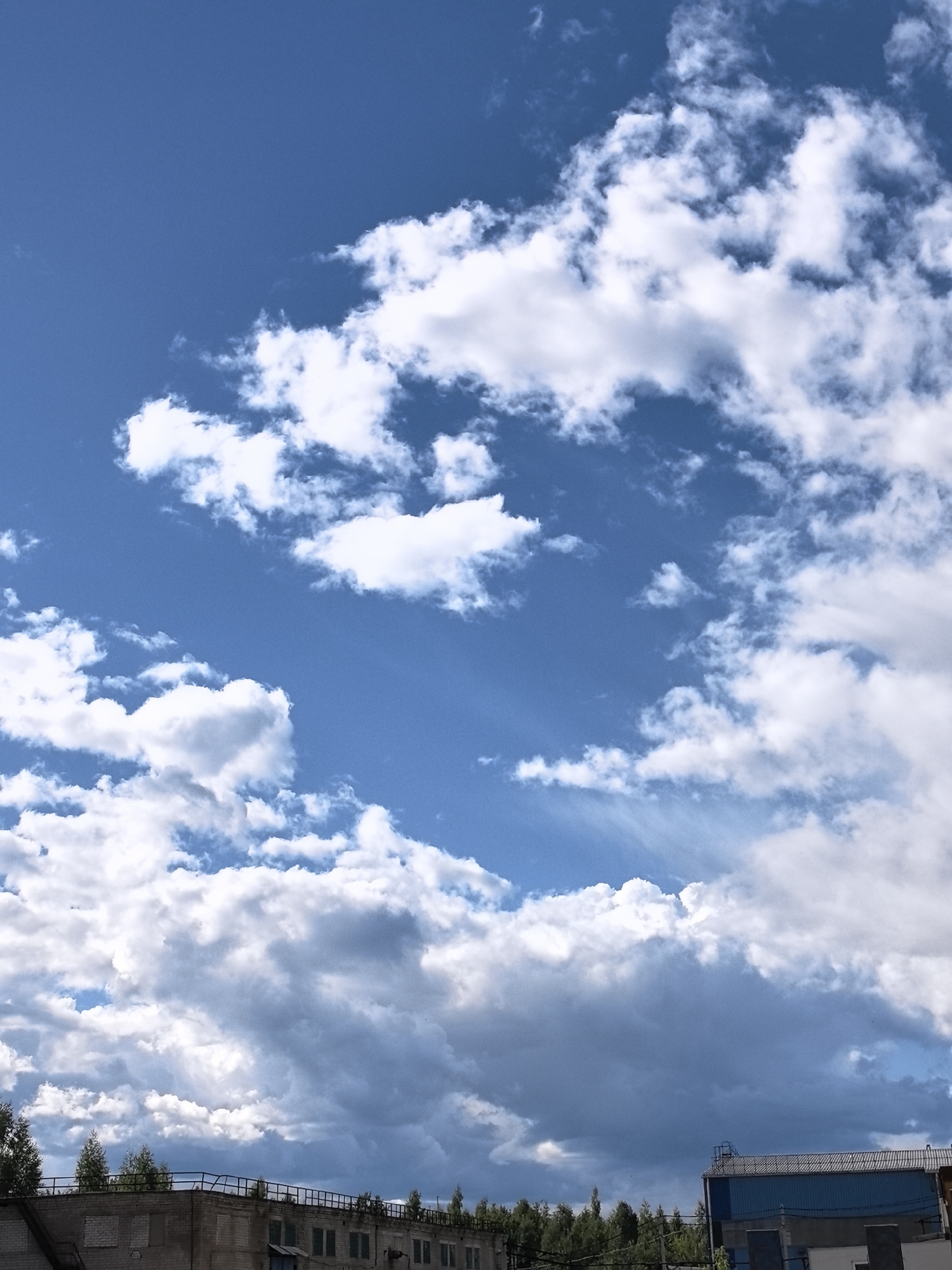 The image shows a bright blue sky filled with fluffy white cumulus clouds. Sunlight filters through the clouds, creating streaks of light in the sky. In the lower part of the image, the tops of buildings and trees are visible, suggesting an urban or industrial setting. The buildings appear to be made of brick and have a somewhat weathered appearance. There is a fence with railings on top of one of the buildings. The overall impression is of a pleasant day with a dynamic sky, likely late morning or early afternoon given the brightness of the sun. The location is identified as Ivanovo, Russia. No people are visible in the image.