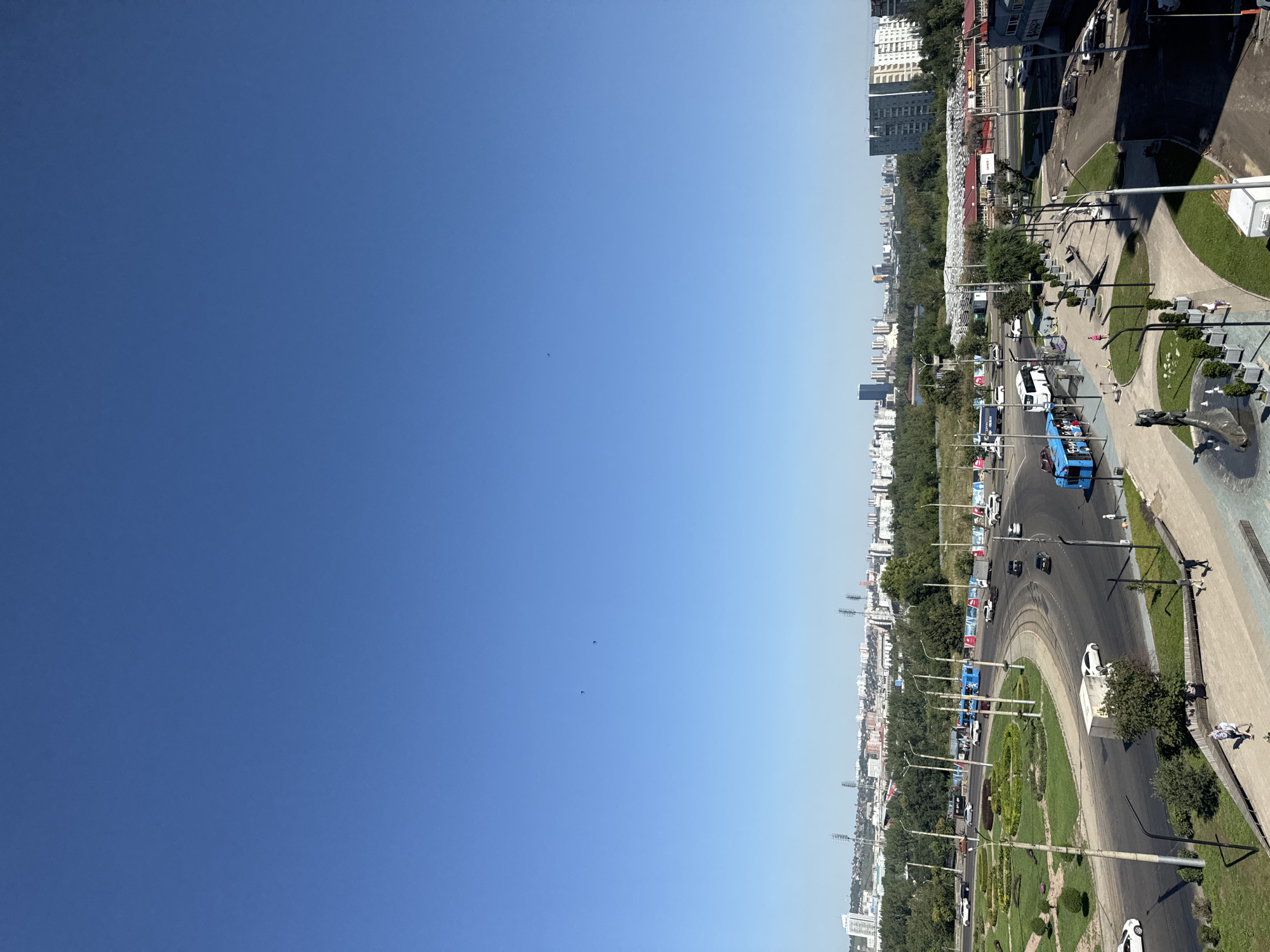 The image is an aerial view of a city street in Krasnoyarsk, Russia, on a clear, sunny day. A blue electric bus and several cars are visible on the road. There are also streetlights, trees, and buildings in the background. In the foreground, there is a pedestrian walkway with benches and a statue. Two people are walking on the walkway. The sky is clear and blue with no clouds.