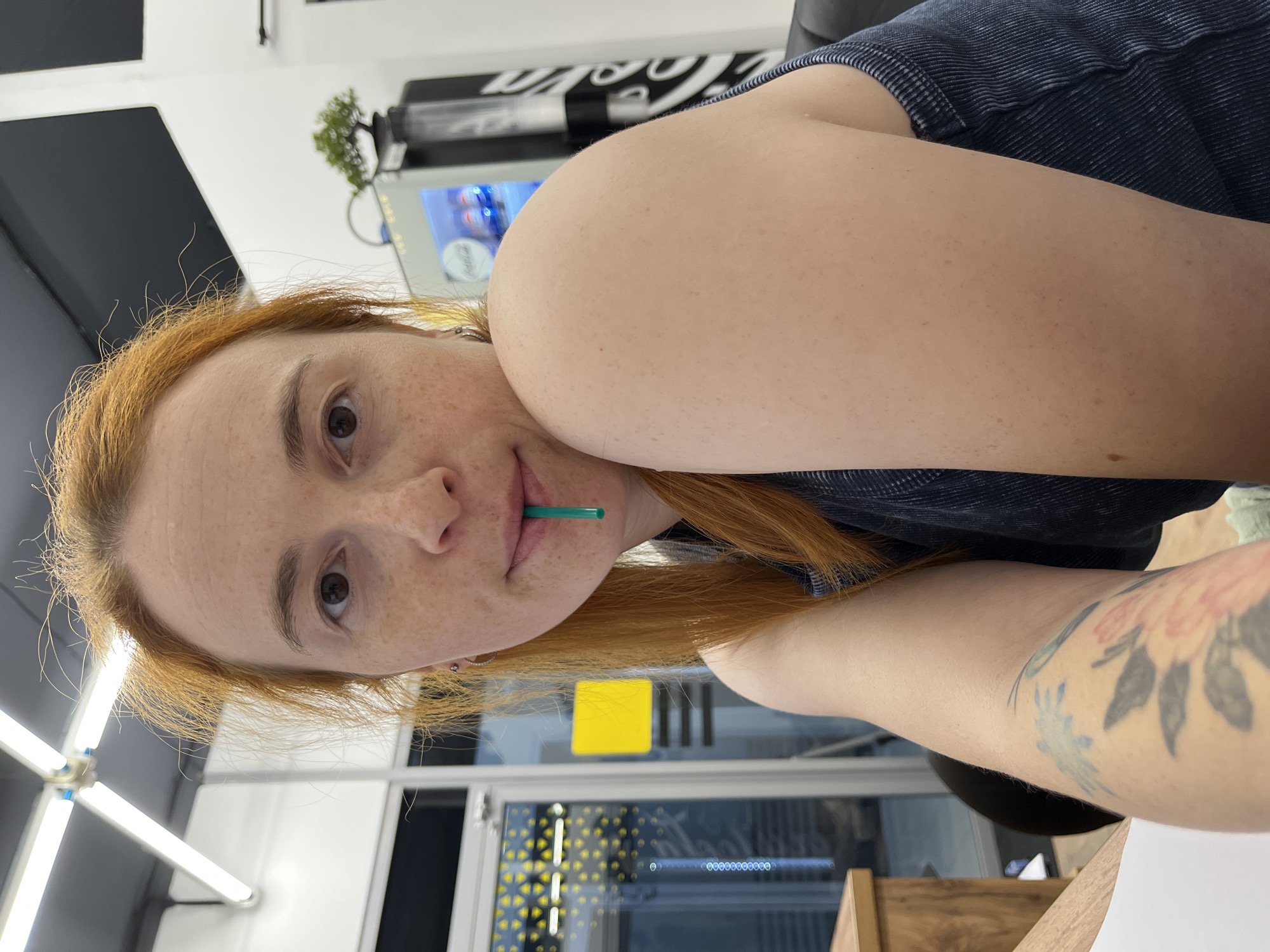 A woman with red hair and freckles is taking a selfie. She has a green straw in her mouth. She is wearing a dark blue tank top. There are tattoos on her arm. In the background, there is a vending machine with Coca-Cola branding and a small bonsai tree. The environment appears to be an indoor space with fluorescent lighting. The image quality is clear and sharp. The location is not specified.