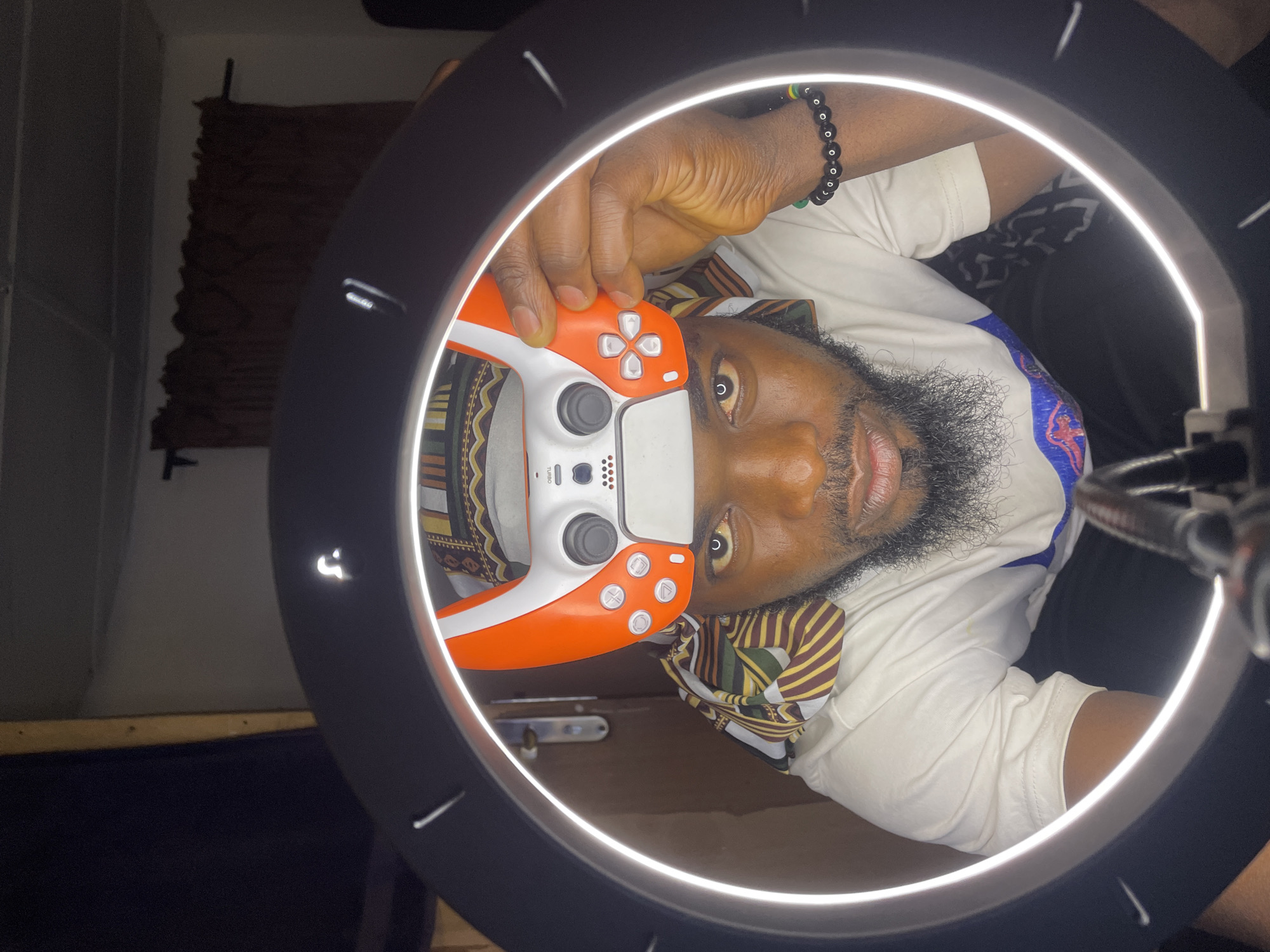 A man with dark skin, a beard, and a bracelet is holding an orange and white gaming controller in front of his face. He is wearing a patterned scarf and a white shirt. The man is positioned within a circular ring light. In the background, there is a dark curtain or tapestry hanging on a wall. The environment appears to be indoors. There is no information to support that the location is Jos, Nigeria.