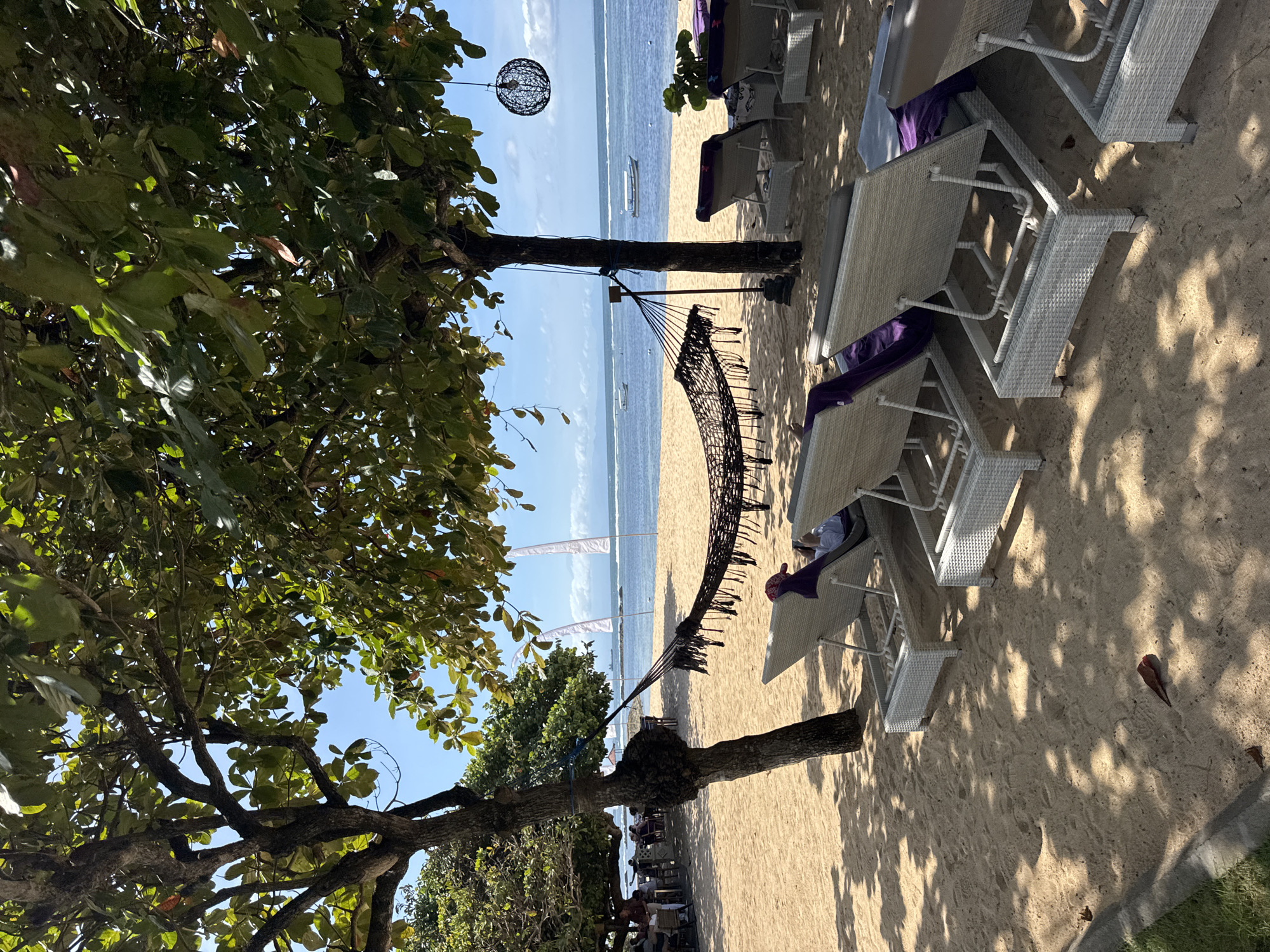 The image depicts a beach scene in Kuta Selatan, Indonesia. A black hammock is suspended between two trees on a sandy beach. To the right of the hammock, there are several white beach lounge chairs with purple cushions. The ocean is visible in the background, with a few boats on the water and white flags on the beach. The sky is mostly clear and blue. To the left, a large tree with green leaves provides shade. People are visible under the trees in the distance. A decorative spherical lamp hangs from one of the trees.