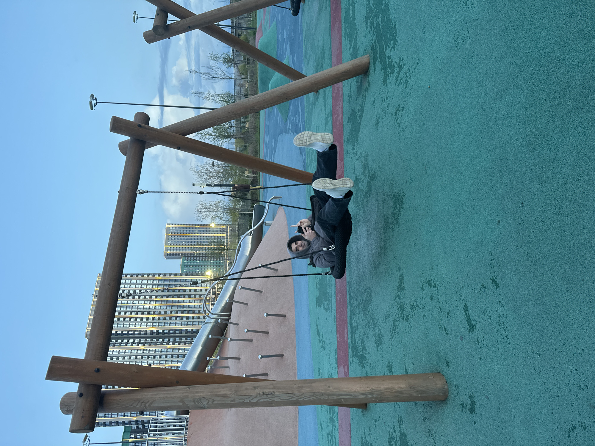 A person wearing a gray hooded jacket, black pants, and white shoes is on a swing at an outdoor playground. The playground has a padded green and red surface, wooden support structures for the swings, and a metal slide with climbing pegs. Tall apartment buildings can be seen in the background. The location is identified as Ovoshchnoy, Russia.