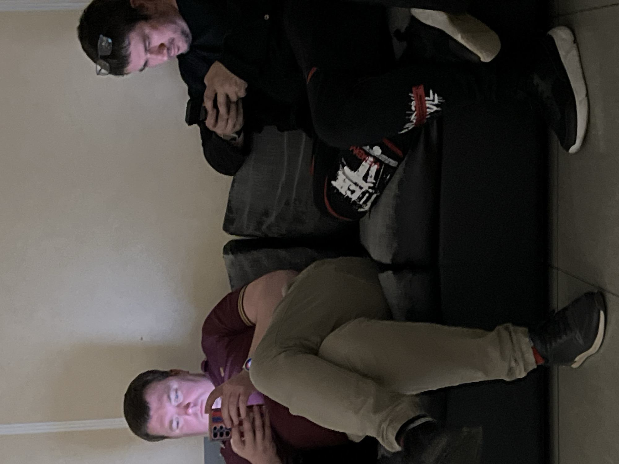 Two men are sitting on a dark-colored sofa. One man is wearing a dark jacket and pants with glasses, and he is looking at his phone. The other man is wearing a burgundy shirt and tan pants, also looking at a phone. The location is not discernible from the image and therefore I cannot identify if this photograph was taken in the city of Khimki, Russia.