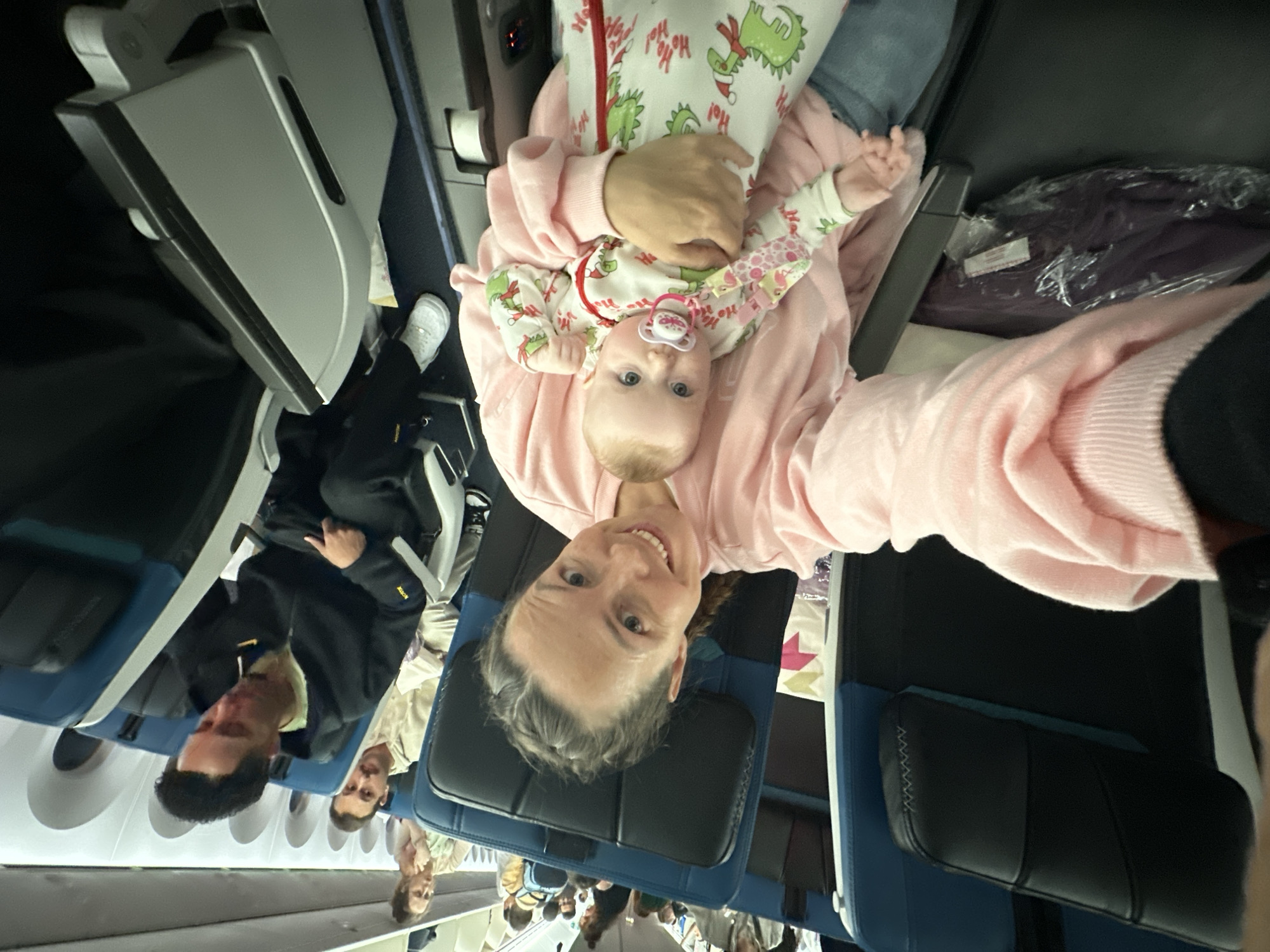 This media file captures a selfie taken inside an airplane cabin, oriented ninety degrees counter-clockwise. The main subjects are a woman in the foreground and a baby she is holding. The woman, with greyish hair, is looking directly at the camera with a smile and is wearing a pink sweater or top. The baby, with blue eyes, is wearing a light-colored onesie adorned with green dinosaur-like figures and the text "Ho Ho Ho!". It has a pink pacifier in its mouth and appears calm and alert.
In the background, several other passengers are visible, seated in various rows. Many appear to be men. The airplane interior features blue and black upholstered seats, white window frames, and grey overhead compartments. A closed tray table is visible on the seat in front of one of the passengers. Under a seat, a clear plastic bag contains what appears to be a folded purple garment. One passenger in the middle distance wears white sneakers. The lighting suggests it is daytime, or the cabin lights are on. The overall scene depicts a moment of travel within a commercial aircraft, possibly during boarding, cruising, or waiting to deplane, with a cheerful interaction captured in the selfie.