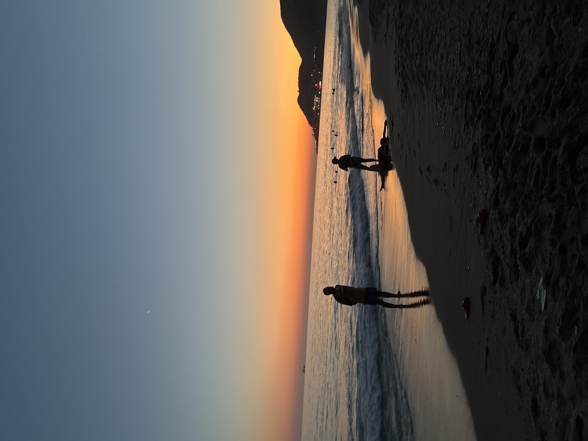 A beach scene in Alanya, Turkey, captures the twilight hours with a vibrant sunset painting the sky with hues of orange and blue. The calm sea gently laps at the shore, reflecting the warm colors of the sunset. Silhouetted figures dot the beach, some wading in the water, others standing by the shoreline. In the foreground, a person lies on the sand while another stands over them. The distant mountains are illuminated by the fading light, with a few lights twinkling on their slopes. A small boat is visible on the horizon, adding to the serene ambiance of the scene. The sand is dark and textured, with some footprints and a few scattered flip-flops. The overall atmosphere is peaceful and tranquil, evoking the beauty of a summer evening.