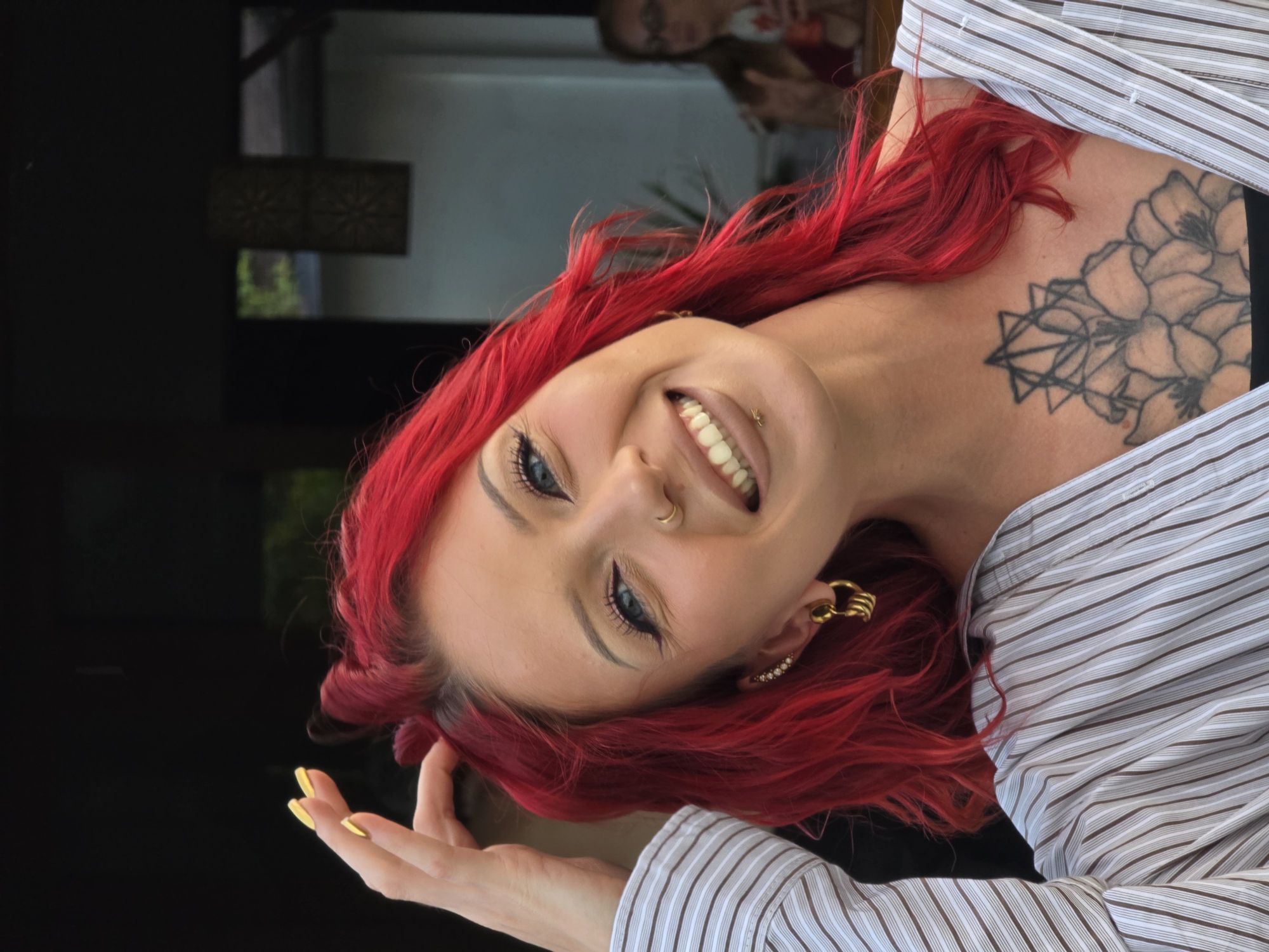A portrait-style image captures a young woman with vibrant, bright red hair and light blue eyes, smiling broadly towards the camera. The image is rotated, presenting her head tilted significantly, as if leaning back or looking up. She has fair skin and is adorned with a gold hoop nose ring and a small stud philtrum piercing. Her smile reveals white teeth. On her upper chest and shoulder, a prominent tattoo features a combination of geometric shapes and floral designs.
She is wearing a light-colored, possibly white, striped button-down shirt with thin brown or black vertical stripes. A hand with bright yellow nail polish is visible, gently touching her hair on the left side of the frame. In her ear, she wears multiple gold-colored earrings, including a large, textured hoop. The background is predominantly dark on the left, transitioning to a reflective surface on the right, which shows a blurred reflection of another person (possibly a woman wearing a red or orange top) and an ornate rectangular light fixture. The setting appears to be indoors, possibly near a window or mirror. The woman's expression conveys happiness and confidence. No text is visible in the image.