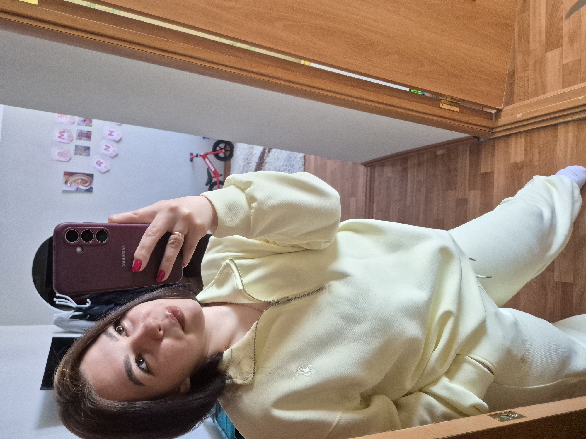 A woman in a light yellow tracksuit is taking a selfie with a burgundy smartphone. She's lying down with a door frame and wooden flooring in the background. There is a white wall with children's artwork and a small red bicycle visible. The location is indicated as Tobolsk, Russia.