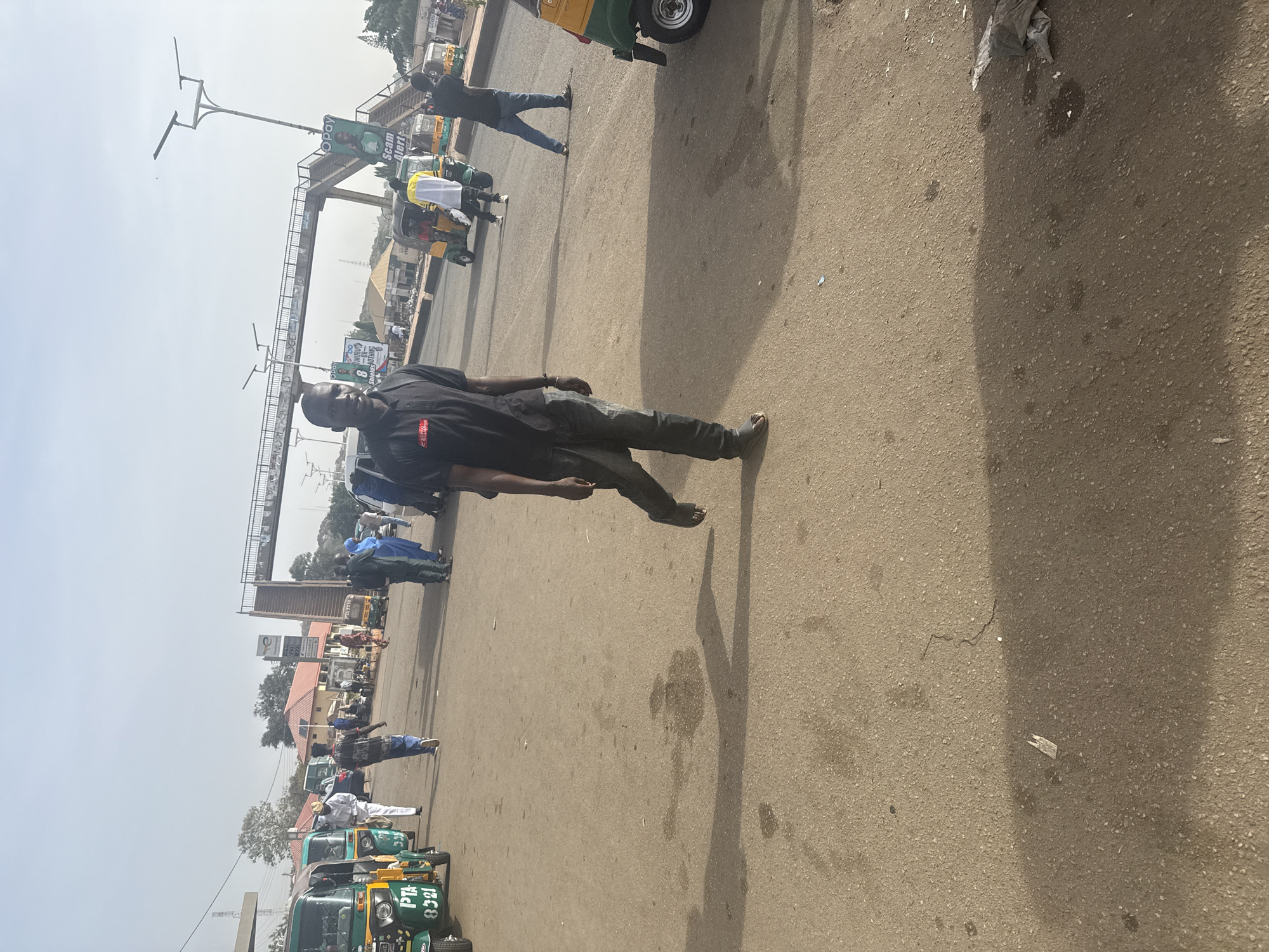 The image depicts an outdoor scene in Jos, Nigeria. A man is walking on a paved surface. He is wearing dark clothing. In the background, there are vehicles, including tuk-tuks, and other people. An elevated pedestrian walkway or bridge is visible in the midground. The sky is overcast.