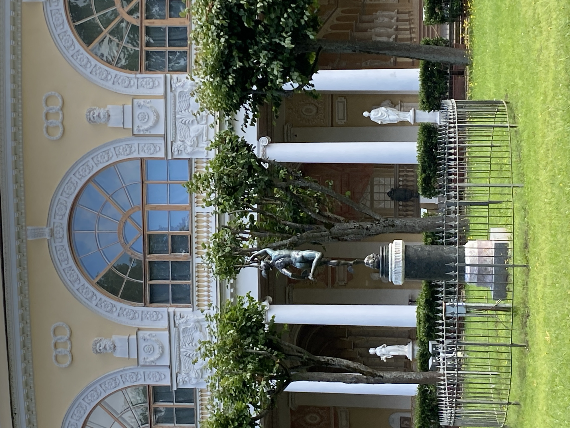 The image captures a view of the Palace of Pavlovsk in Pavlovsk, Russia. In the foreground, a circular black metal fence encloses a grassy area with a bronze statue of a nude male figure on a dark pedestal. Behind the fence, on the left, a white marble statue of a standing female figure is visible. To the right of the bronze statue, another white marble statue of a standing female figure is partially obscured by a tree.
The background features a pale yellow building with a classical facade. The building has large arched windows with a radiating pattern, separated by ornate white stucco decorations including busts of men and winged mythical creatures. White columns support an upper level with a balustrade. Green trees with full canopies are strategically placed throughout the scene, framing the architectural elements and statues. The overall setting suggests a well-maintained historical park or garden.