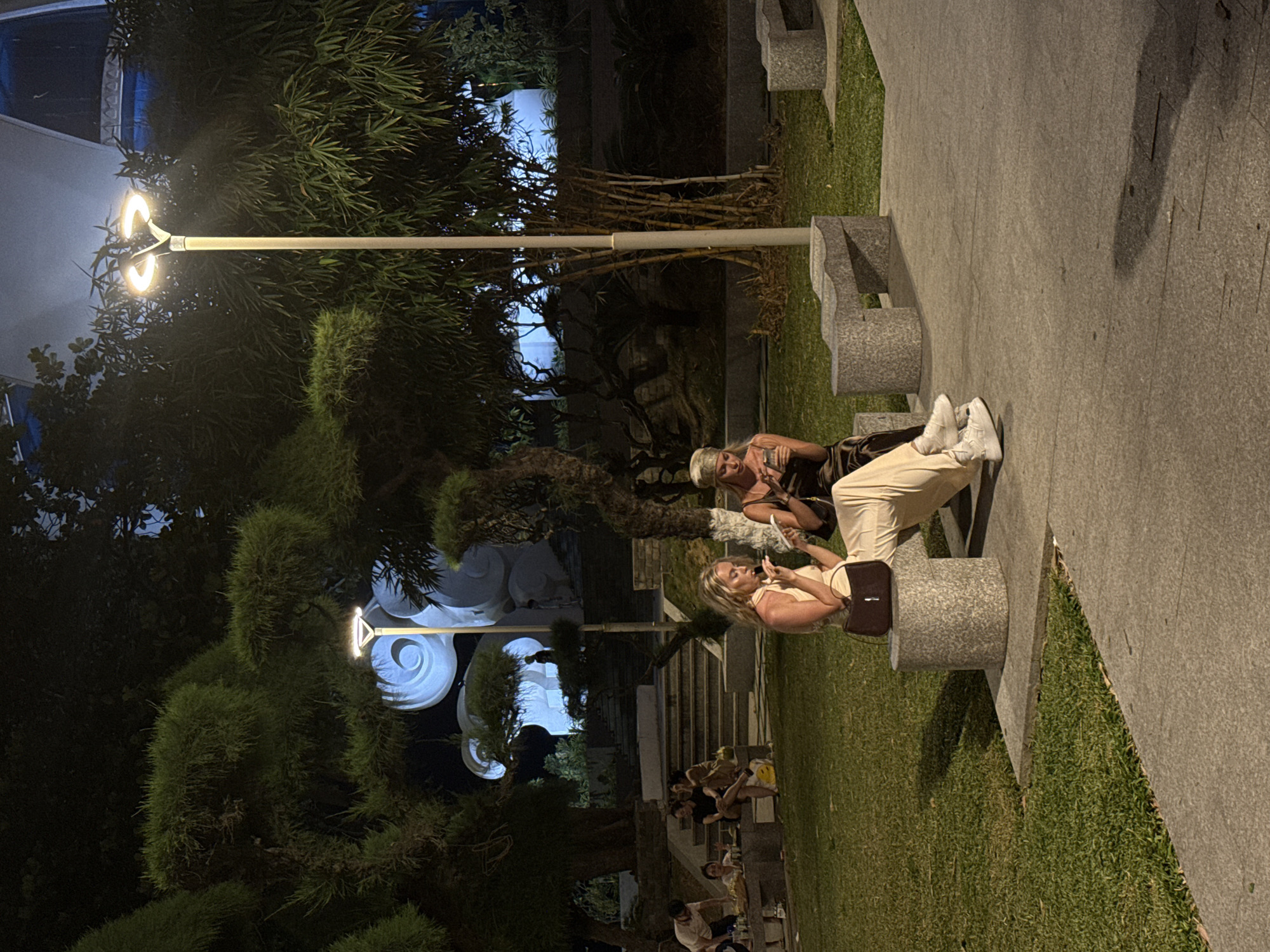 The photograph shows an outdoor scene in Nha Trang Ward, Vietnam, featuring benches along a walkway adjacent to a grassy area. Two women are seated on a bench; one is holding and looking at a mobile phone. Other people are sitting on steps in the background. A street light illuminates the scene. Trees and ornamental structures provide background detail.