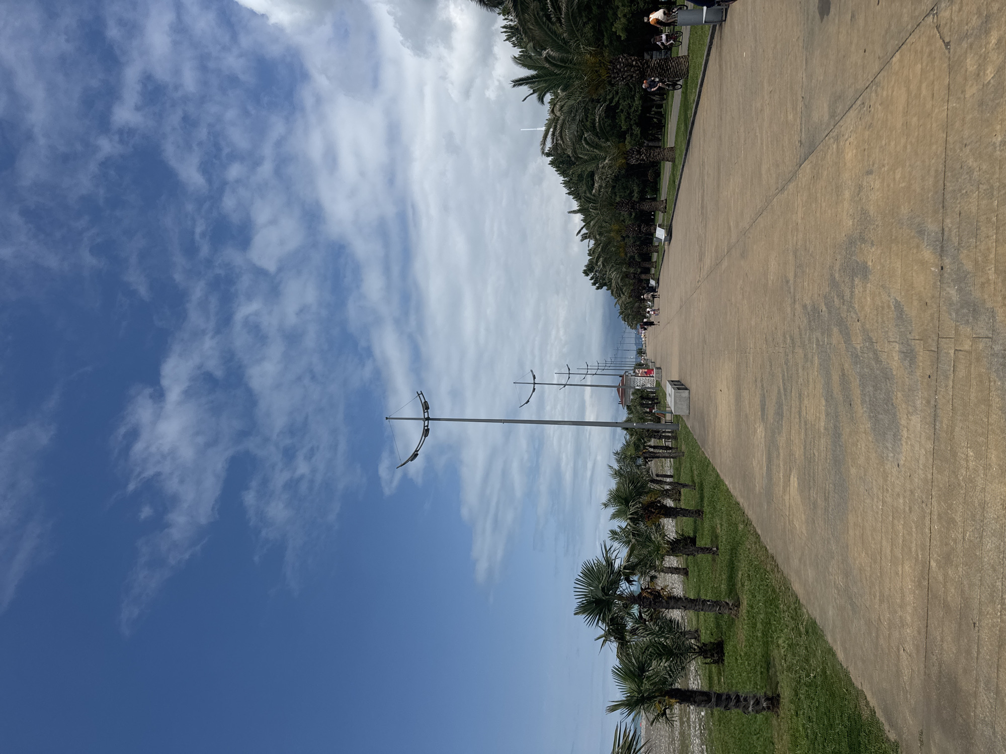 The image depicts a wide, paved promenade adjacent to a lush green lawn lined with palm trees, leading towards a densely wooded area. The promenade is busy with people walking, cycling, and one person on a scooter. The sky above is a bright blue, dotted with scattered white clouds, suggesting fair weather. Several modern lampposts with unique, curved designs are spaced along the promenade. In the background, buildings and a tower can be seen peeking through the trees. To the left of the promenade, there is a glimpse of a pebbled beach and the sea. The scene is likely the Batumi Boulevard, a popular waterfront promenade in Batumi, Georgia, known for its palm trees and recreational spaces.