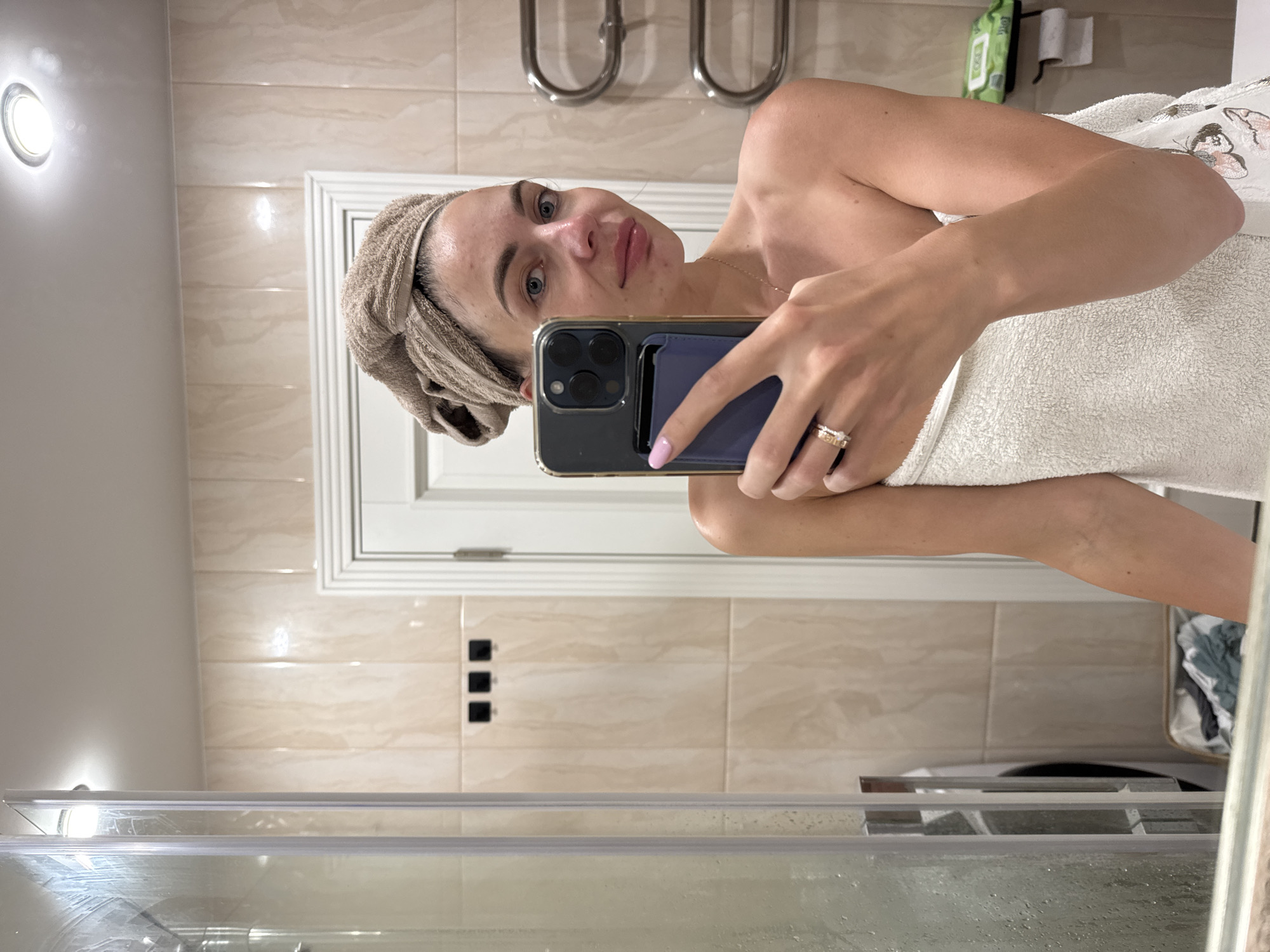 A young woman with a towel wrapped around her head and body takes a selfie in a bathroom. She is holding a smartphone in her right hand, angled to capture her reflection in a mirror. The bathroom features tiled walls, a shower enclosure, a towel rack, and a washing machine visible in the background. There are also some wet wipes and toilet paper on a shelf. The image appears to be taken in a domestic setting. The location is not specified in the image.