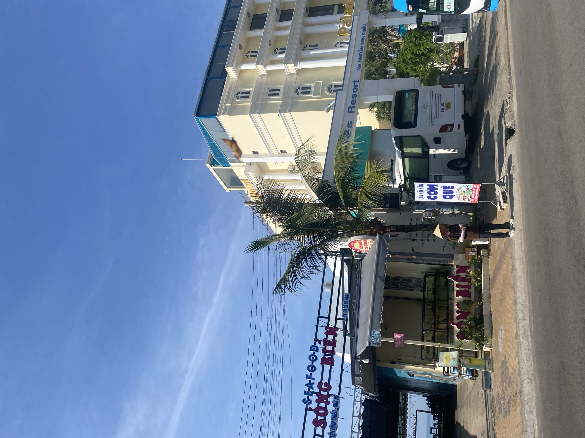 The image shows a street scene in Phu Thuy Ward, Vietnam, with a seafood restaurant and a resort. On the left, a sign for "SEAFOOD SÔNG BIÊN" is visible. A man wearing a conical hat stands on the sidewalk in front of the restaurant, which has several fish tanks displayed outside. To the right of the restaurant, a white bus is parked, and a sign for "LẨU HẢI SẢN CƠM QUÊ" is placed in front of it. Further to the right, a large yellow building with white columns, identified as "Paladin Resort", dominates the background. A blue bus is partially visible on the far right. The sky is clear and blue.