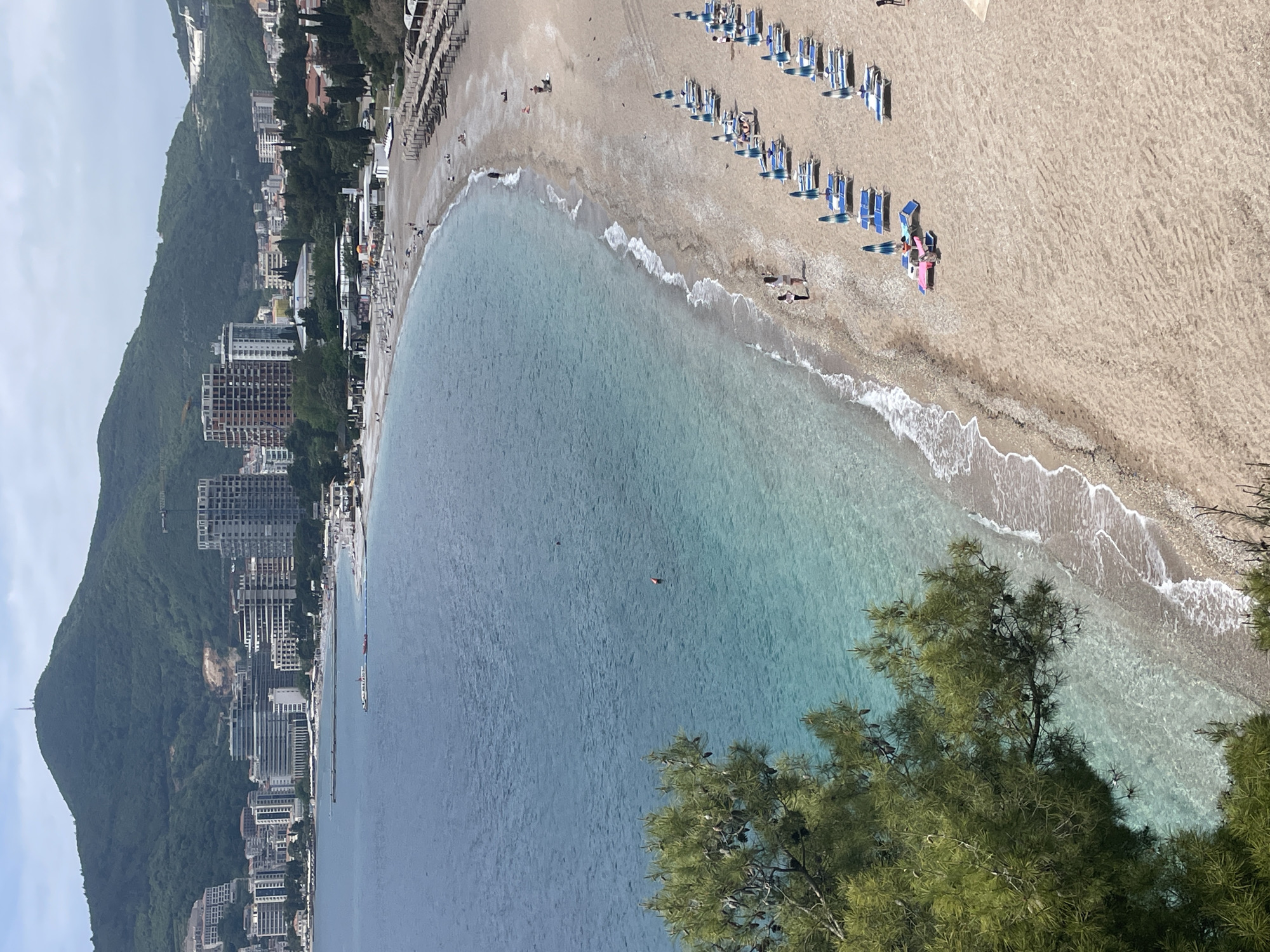 The image shows a beach in Budva, Montenegro, from an elevated perspective. The beach is sandy with rows of sunbeds and umbrellas. The water is clear turquoise. Buildings and green hills are on the left side of the frame. A single swimmer is visible in the water. Tree branches are in the lower right corner.