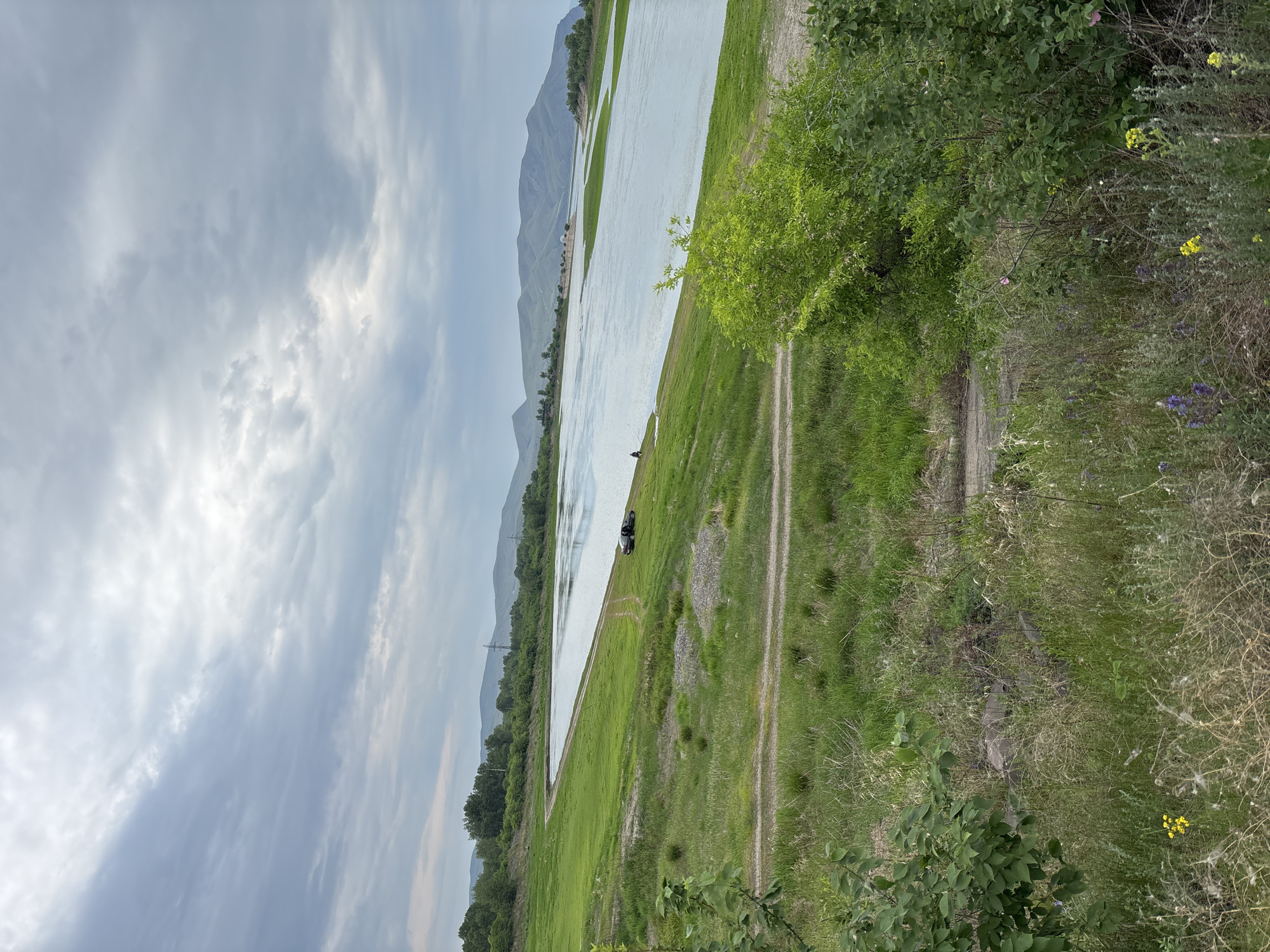 The image depicts a wide, calm river flowing through a green, grassy landscape under a cloudy sky. The riverbank is lined with lush green grass and vegetation. In the distance, rolling hills and mountains are visible. On the grassy bank near the river, a dark-colored car is parked. A dirt path or road can be seen winding along the riverbank. The overall scene suggests a natural, possibly rural or semi-rural setting near Ust-Abakan, Russia.