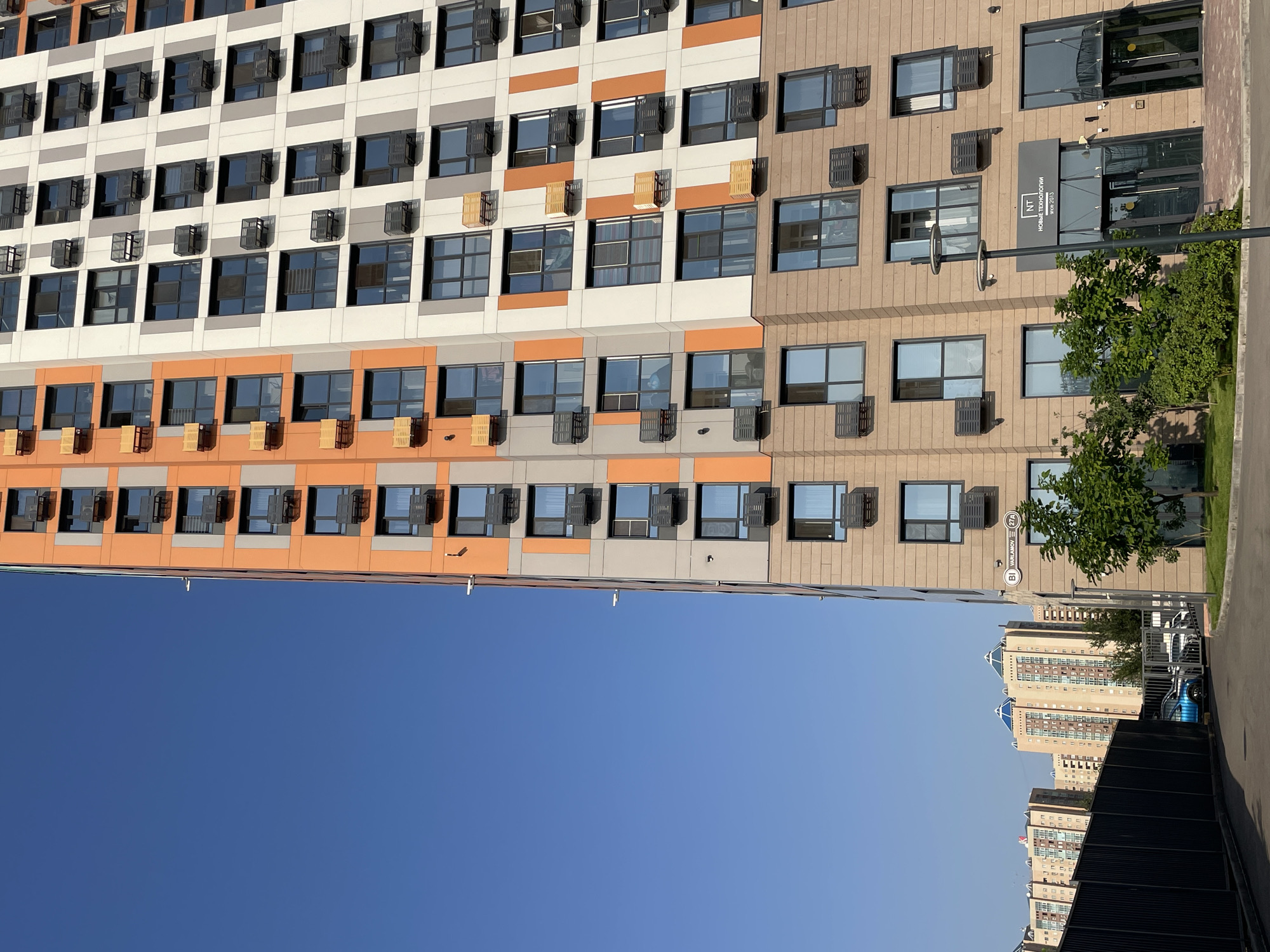 The image captures the facade of a modern, multi-story apartment building in Almaty, Kazakhstan, under a clear blue sky. The building features a striking color palette with sections of orange, gray, and beige. Numerous windows are visible, many equipped with dark-colored air conditioning units. Some windows on the orange sections also have lighter colored frames that appear to be for balconies or window boxes. In the foreground, a dark fence lines a paved area, with a blue car and other vehicles visible beyond it. To the right, the entrance to the building is visible, featuring large glass doors and a sign that reads "NT НОВЫЕ ТЕХНОЛОГИИ since 2013". A street lamp stands in front of the entrance, and lush green trees and bushes are planted in front of the building. In the background, other apartment buildings, some with distinct blue pyramid-shaped roofs, are visible.