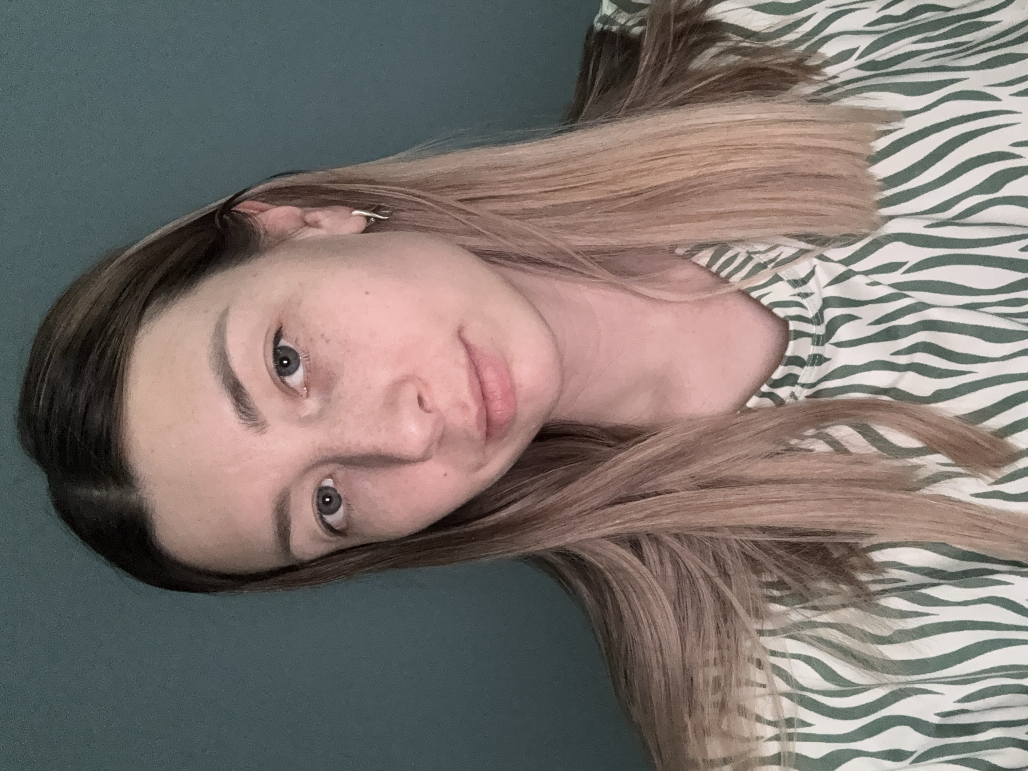 A young woman with light skin, freckles, and light brown hair with blonde highlights is shown from the shoulders up. She is wearing a white shirt with green zebra stripes. A simple silver earring is visible in her left ear. The background is a solid teal color. The setting is not Moscow, Russia.