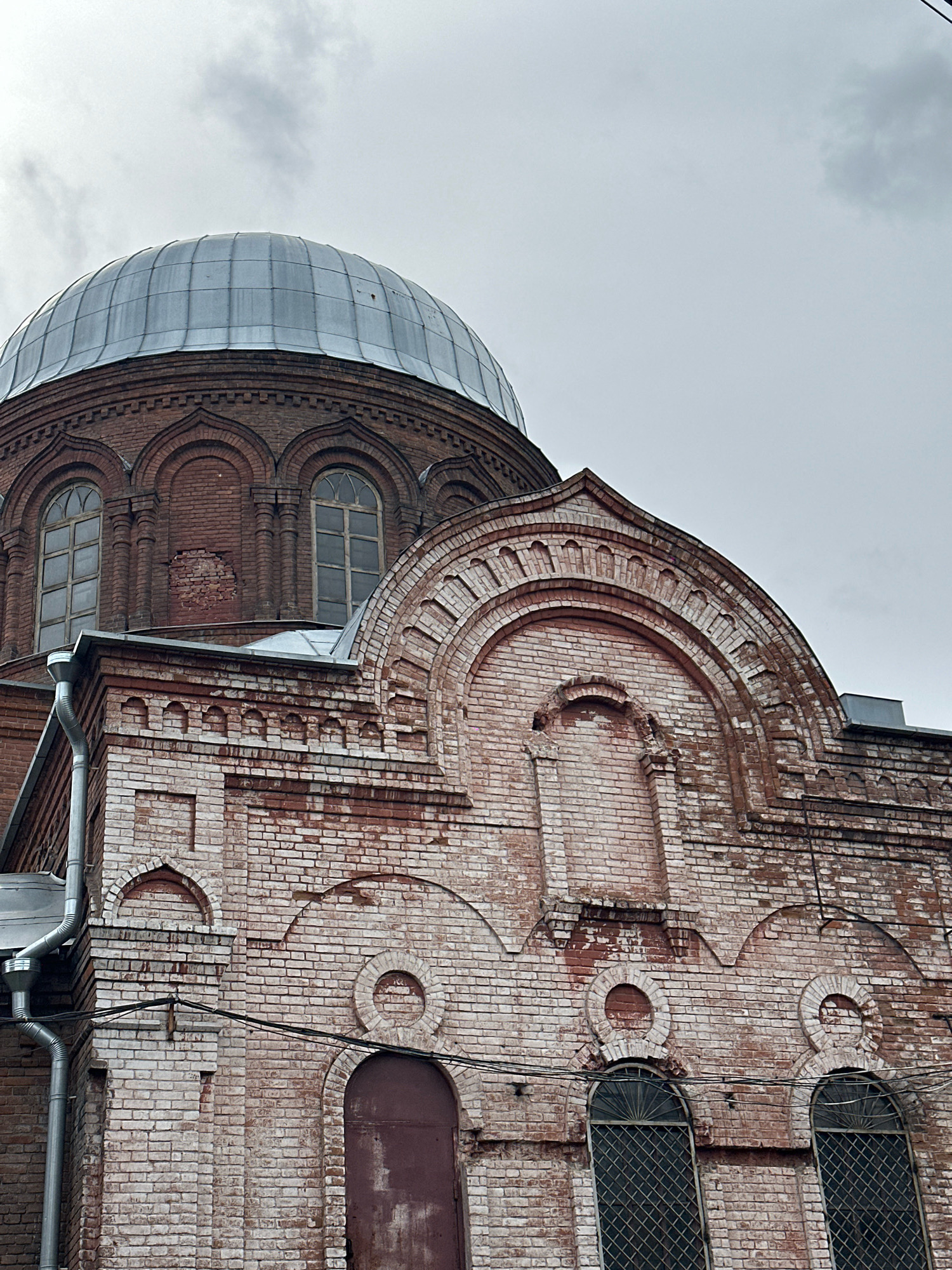 The image shows the exterior of a red brick building with a large domed roof, likely a church or religious building, in Biysk, Russia. The dome is covered in metal panels. The brickwork of the building is ornate, featuring arched windows, decorative brick patterns, and some areas of wear and tear, indicating age. A downspout runs along the side of the building. The sky is overcast, suggesting a cloudy day. There are no people or vehicles visible in the frame. The building appears to be situated in an urban environment, given the presence of overhead wires.