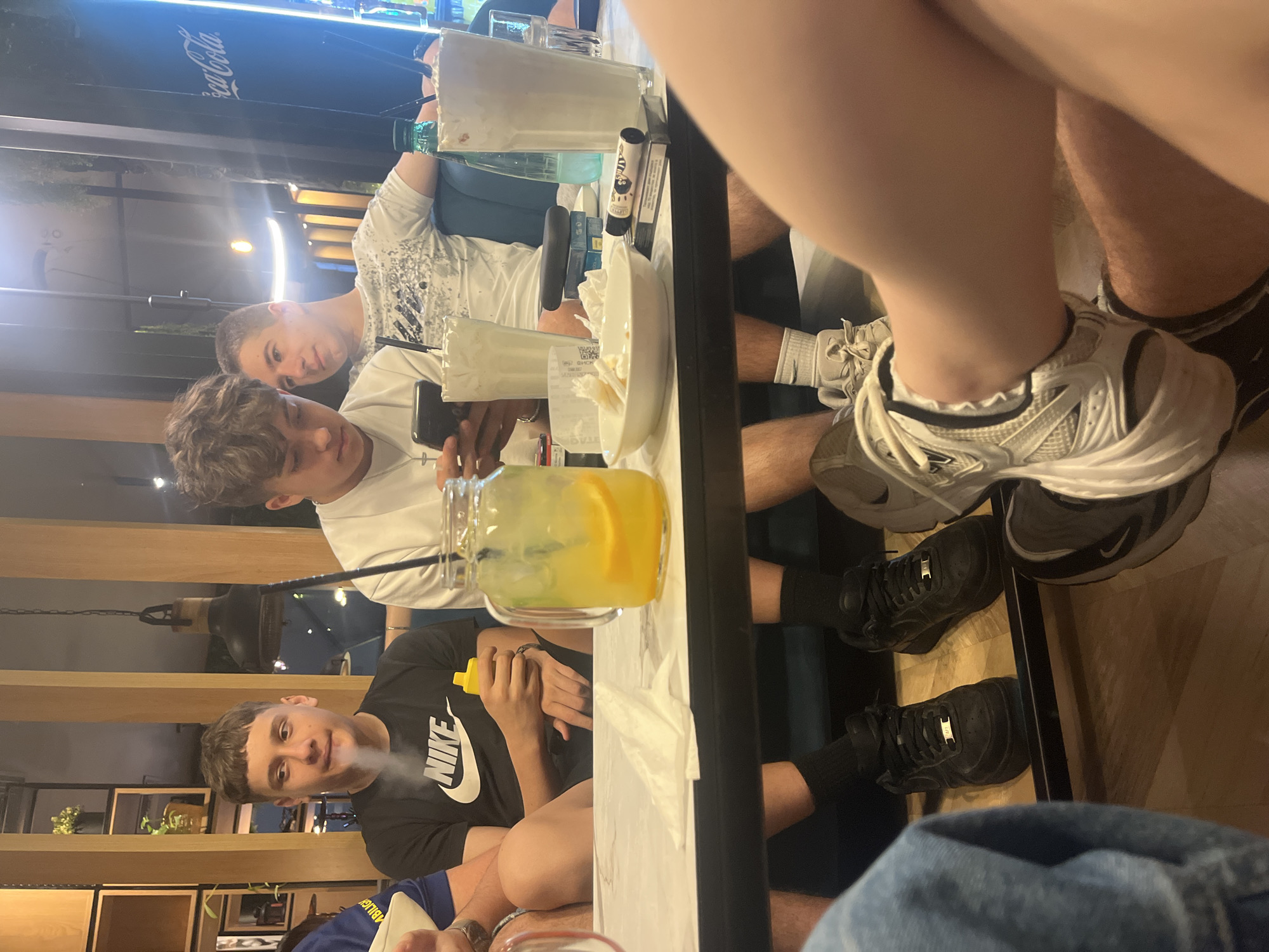 A group of young men are seated around a table at what appears to be an indoor dining establishment. The location is possibly in Stara Zagora, Bulgaria, but this is not definitively confirmed by the visual evidence. The table is a long, rectangular shape with a white surface, and it has various items placed on it, including beverages in glass containers, a plate with food remnants, tissues, and a mobile phone.
The men are dressed casually; one wears a black t-shirt with a white "NIKE" logo, another wears a white t-shirt, and there are glimpses of clothing from other individuals at the table. One of the men is exhaling vapor or smoke. Some are looking at the camera. Their expressions are mostly neutral.
The restaurant has a modern interior with wooden accents and shelving in the background. The lighting appears to be a mix of natural and artificial. A Coca-Cola sign is visible in the background.