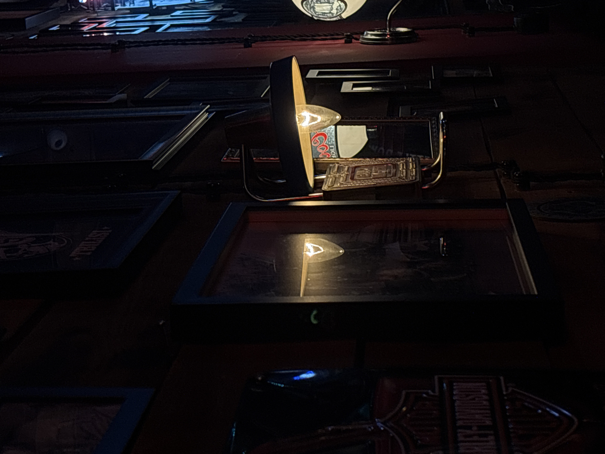 The image shows an interior view of a dark room. A decorative light fixture with an exposed bulb illuminates a collection of framed items mounted on a wall. One frame contains an image of a Coors beer can, while another features a Harley-Davidson logo. The location is unspecified but can be assumed to be in Belgrade, Serbia.