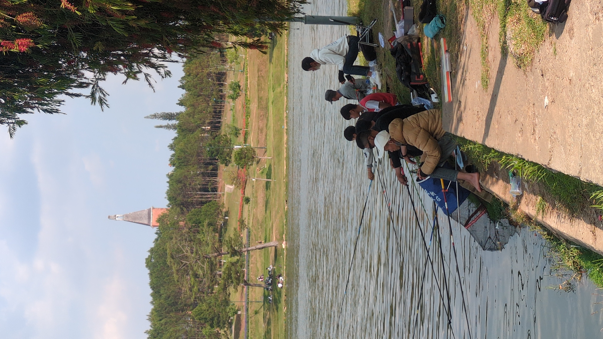 The image shows a group of six individuals fishing at Ho Xuan Huong Lake in Da Lat, Vietnam. The group is lined up along the concrete edge of the lake, each holding a fishing rod. Trees are visible in the background, including the tower of the Da Lat Cathedral on the left. One individual is barefoot. Fishing gear is visible next to them.