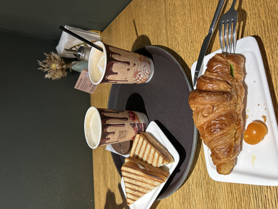 The scene is an indoor cafe setting in Voskresensk, Russia, presented on a light brown wooden table. A dark brown circular tray occupies the midground, holding two disposable paper cups patterned with chocolate drips. Both cups contain a light brown beverage, likely coffee; one cup has a black straw inserted, and a separate brown cup lid rests near the other cup. Adjacent on the tray, a white rectangular plate holds two triangular grilled sandwiches with visible grill marks. In the foreground, a second white rectangular plate features a golden-brown croissant with a green filling visible at one end, alongside a single orange cherry tomato. A silver fork is partially embedded in the croissant, and a silver knife lies parallel to it on the plate. The background consists of a dark green wall. Tabletop decor behind the tray includes a small vase containing dried flowers or botanicals, a silver sugar dispenser, a white napkin holder with white paper napkins, and a small, light-colored rectangular informational card. No individuals are visible.