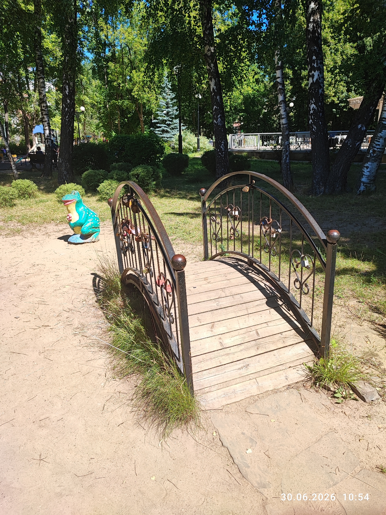The image shows a small, decorative pedestrian bridge made of wood and wrought iron in a park setting. The bridge has a curved design with ornate metal railings that are decorated with heart-shaped elements and several padlocks, suggesting it is a popular spot for couples. The bridge spans a sandy path. To the left of the bridge, a turquoise garden sculpture resembling a dinosaur or frog is situated on the sandy ground. The park is surrounded by lush green trees with visible birch trees among them, indicating a natural environment. In the background, there are hints of other park amenities like benches and a fence. The lighting suggests it is a sunny day. The bottom right corner of the image displays a timestamp reading "30.06.2026 10:54". The scene is consistent with a public park in Lytkarino, Russia, likely a children's play area or a scenic spot within a larger park. There are no visible people in the immediate vicinity of the bridge and sculpture.